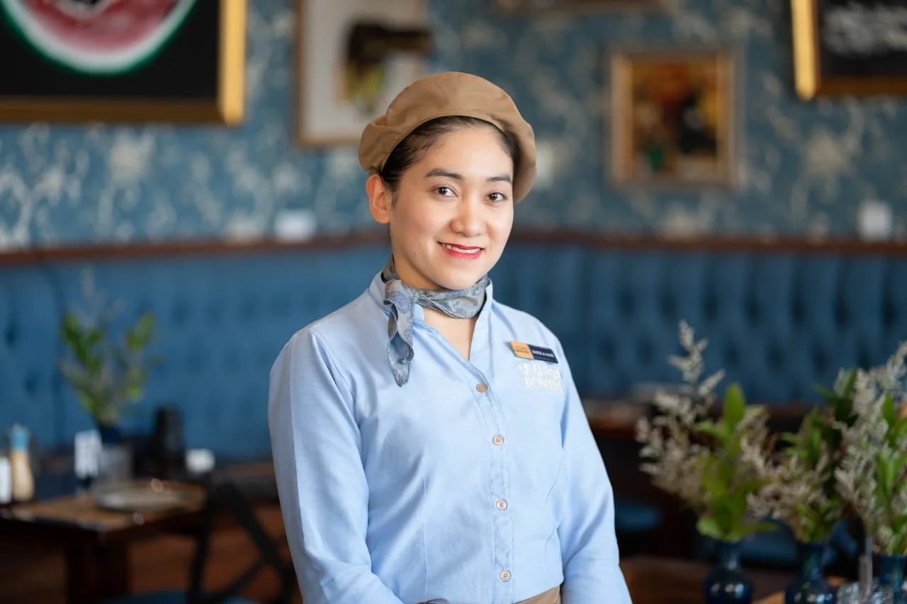 A young woman with dark hair, wearing a light blue server uniform, a tan beret, and a gray scarf, stands inside a restaurant with blue and gold decor. She is smiling at the camera.