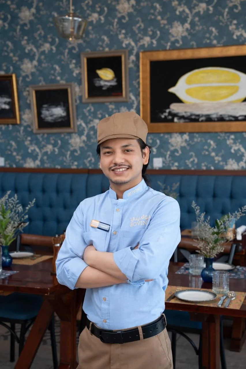 A smiling waiter wearing a beige cap and light blue shirt with his arms crossed, standing inside a restaurant with floral wallpaper, framed artwork, and a dining table set with plates and flowers.