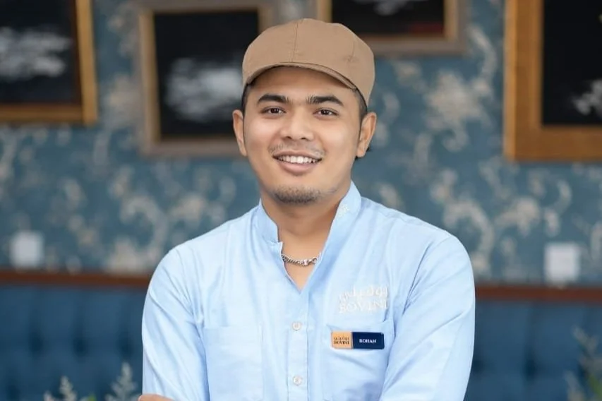A smiling man wearing a light blue shirt, tan cap, and a name tag, standing in a restaurant with blue patterned walls and framed pictures in the background.