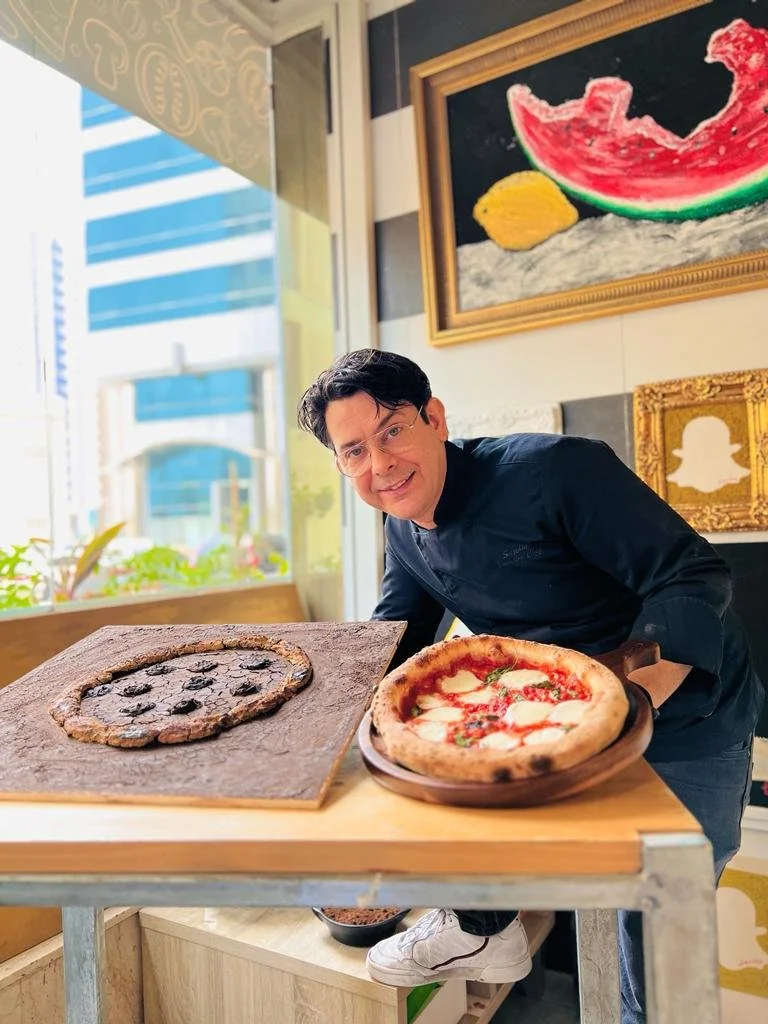 A man in a black chef jacket stands at a counter with two pizzas: one with a black crust and toppings on a stone slab, and another traditional pizza with cheese and pepperoni on a wooden board. Behind him, colorful framed artwork is visible.