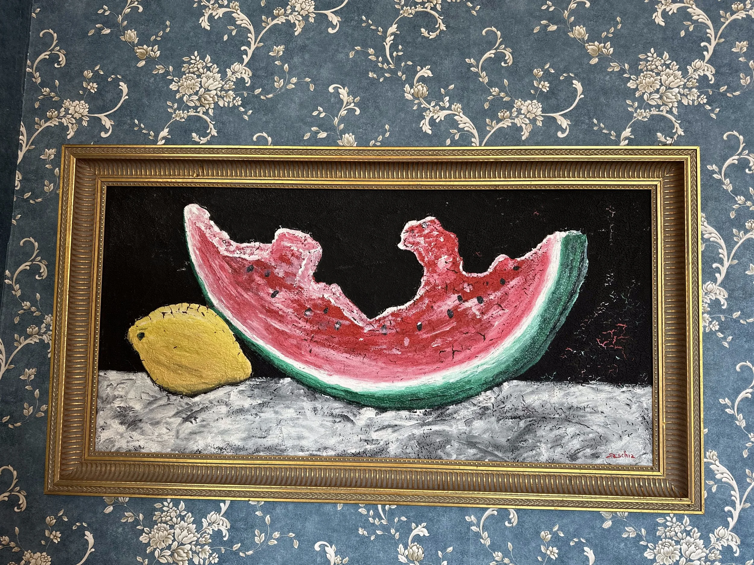 Painting of a watermelon slice and a lemon on a textured surface, framed in an ornate gold frame, against a blue floral wallpaper background.