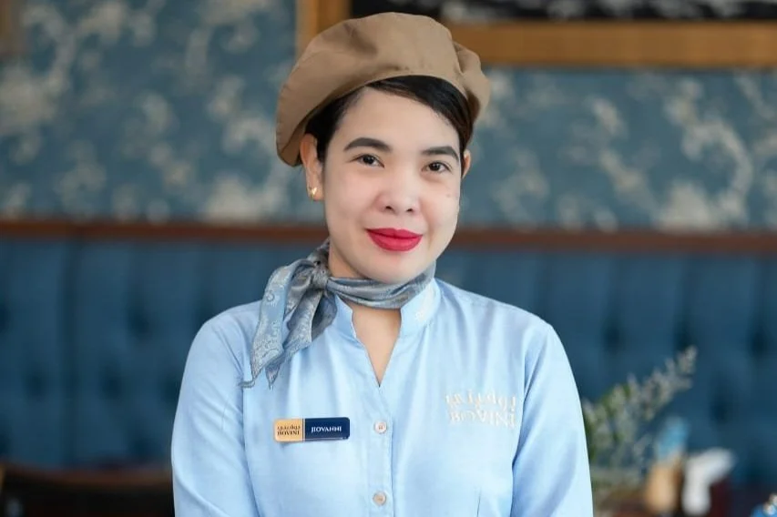 A woman dressed as a hotel or service worker, wearing a tan beret, blue scarf, light blue button-up shirt with a name tag, standing in a restaurant or café with a blue upholstered wall and floral decoration in the background.