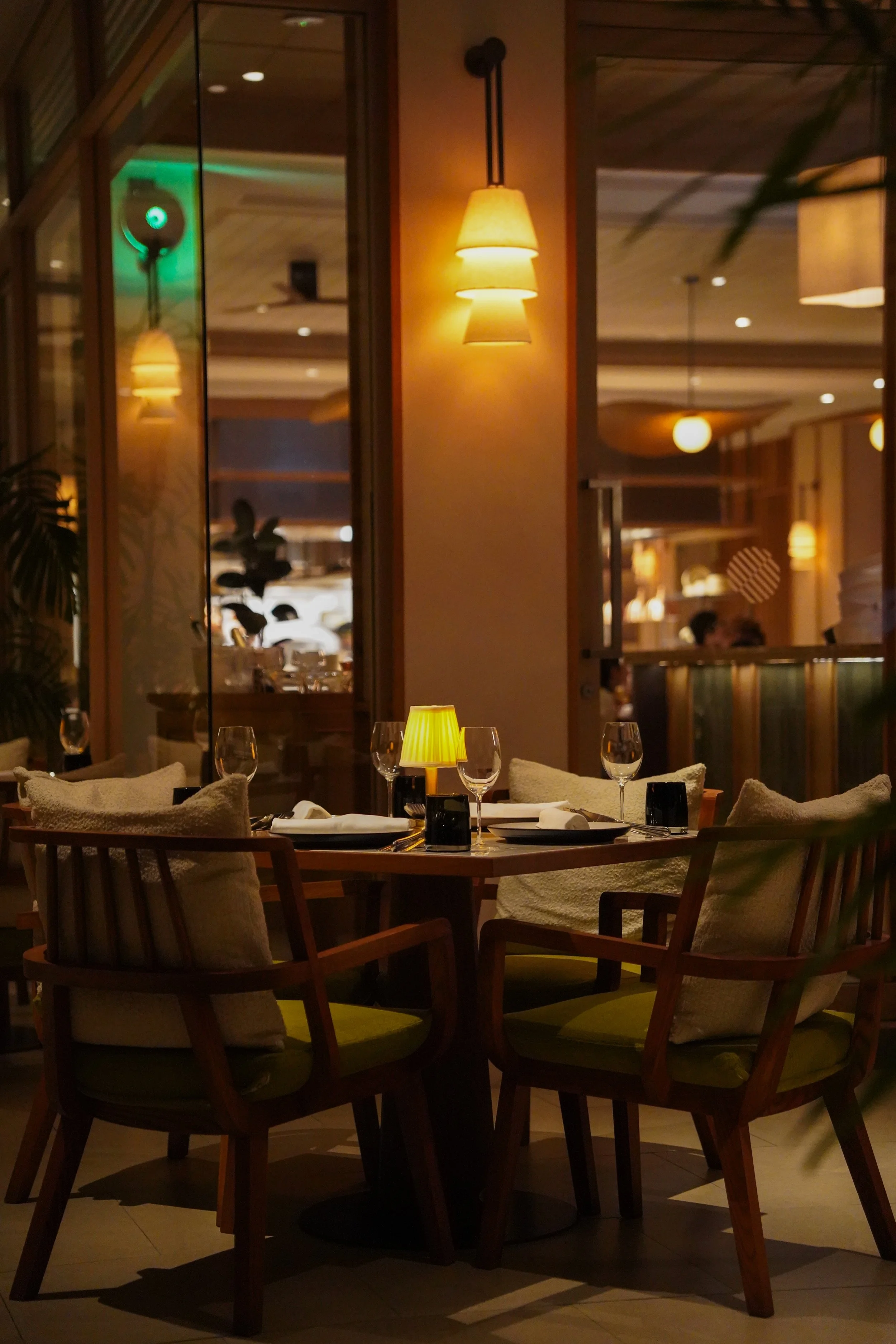 Elegant restaurant table set with wine glasses, cloth napkins, and a small yellow lamp, surrounded by wooden chairs with cushions, in a warmly lit dining area.
