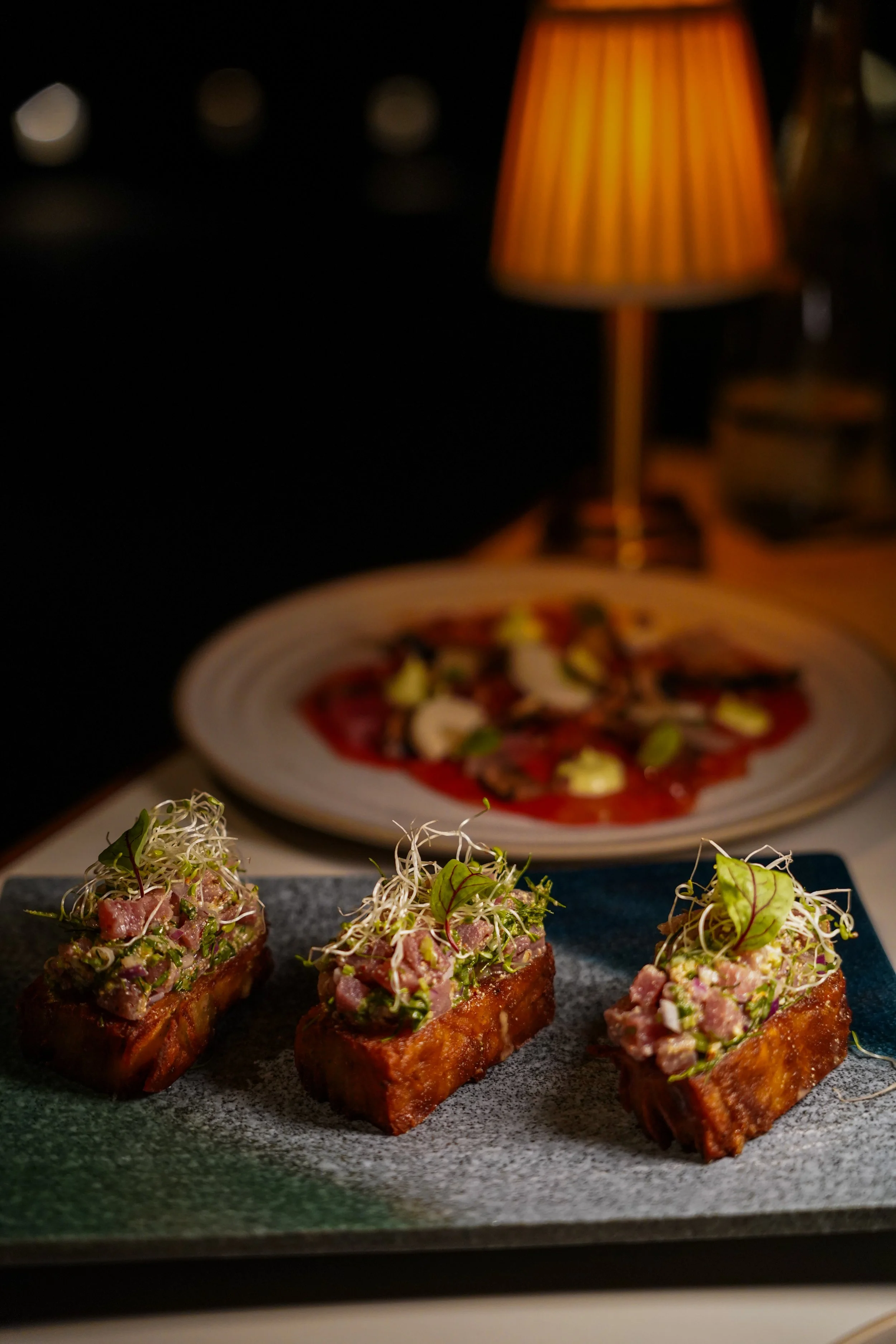Three pieces of tuna poke topped with microgreens, served on a rectangular plate, with a pizza topped with mushrooms and cheese in the background, in a dimly lit setting.