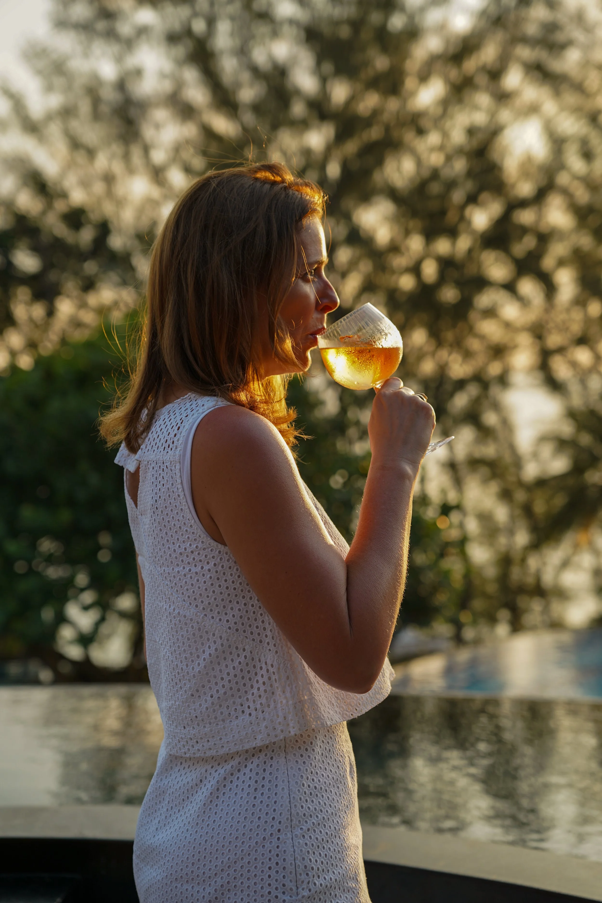 A woman in a white sleeveless dress holding a glass of white wine outdoors at sunset with trees in the background.