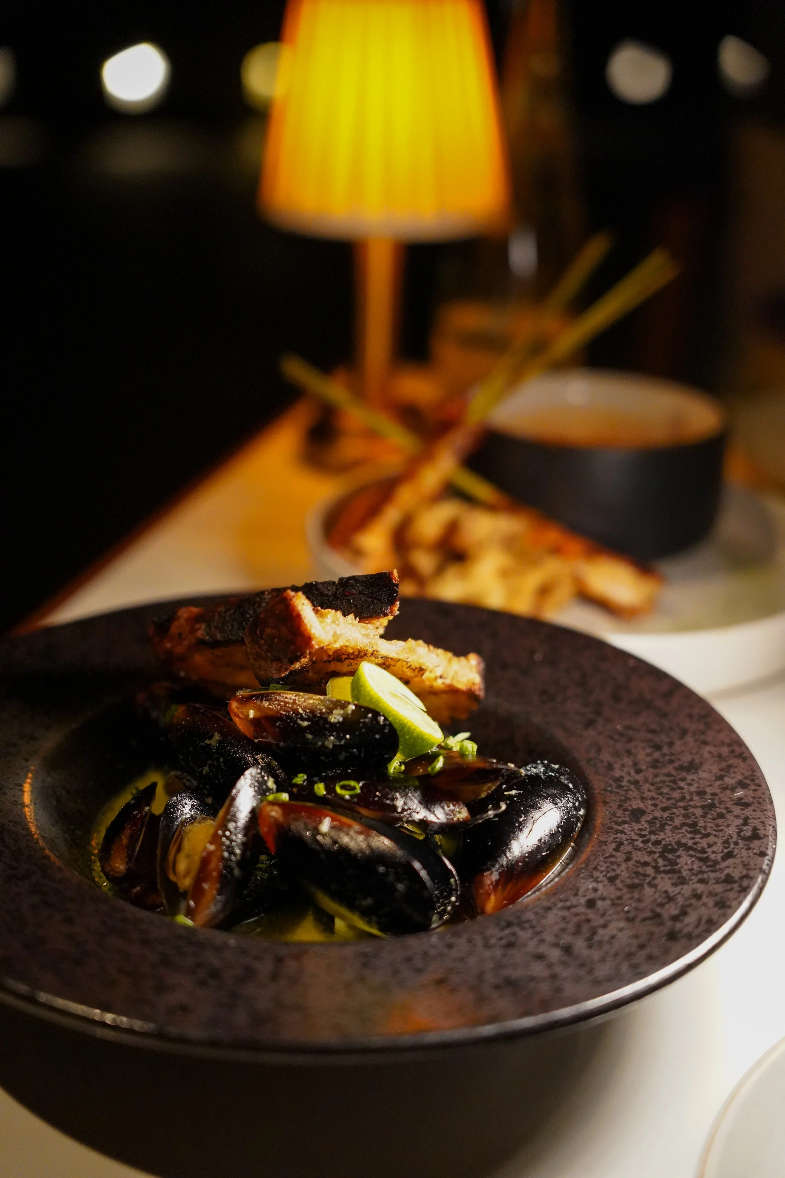 Bowl of mussels garnished with lime and herbs, with toasted bread slices on a black plate, in a dimly lit restaurant setting.