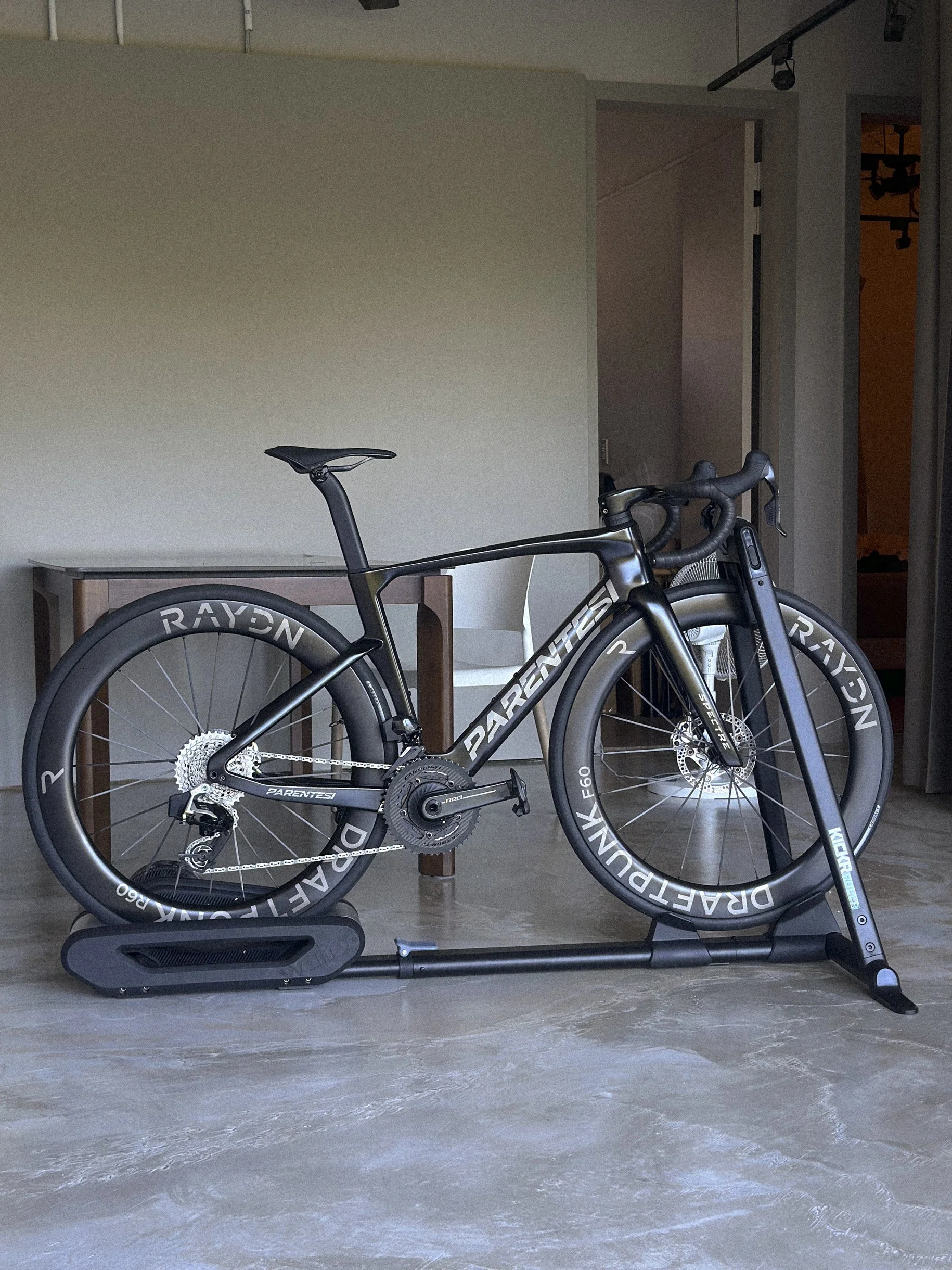 A black PARENTESI bike on an indoor bicycle trainer, positioned indoors on a tiled floor, with a wooden table and white chair in the background.