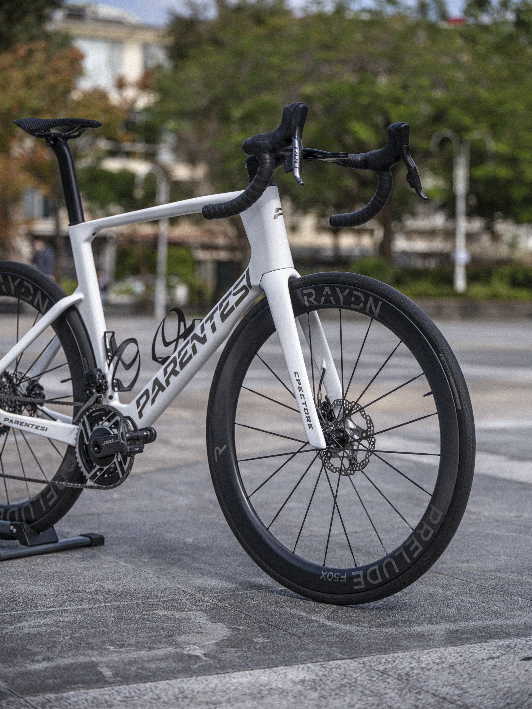 A white Parentesi road bike with black wheels and handlebars, placed on a skateboard rack on a paved area with trees and buildings in the background.