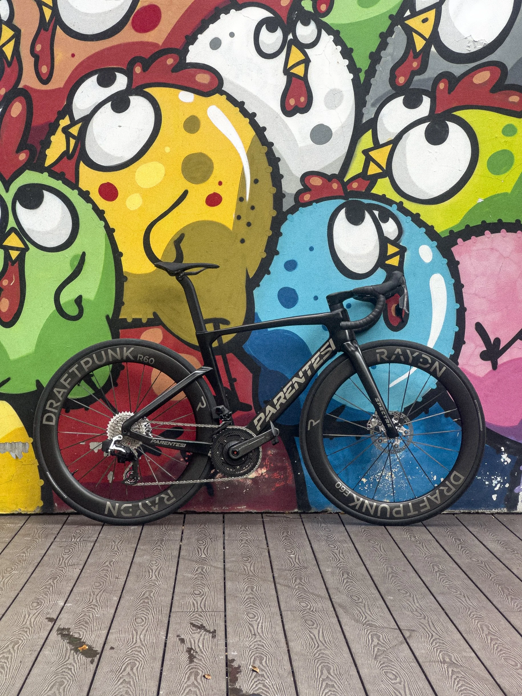 A black PARENTESI racing bicycle in front of a colorful mural of cartoon chickens with large eyes and bright colors.