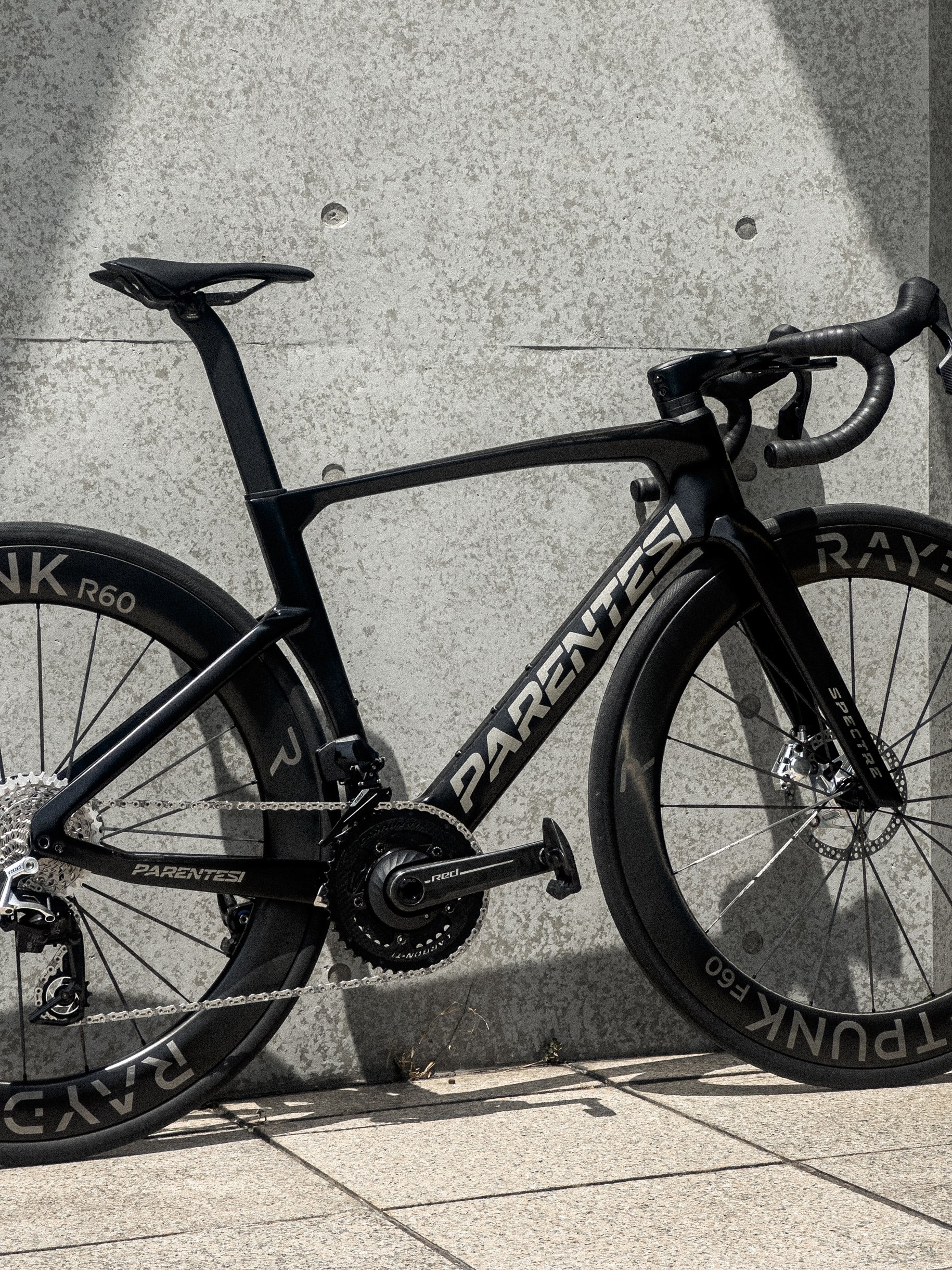 A black PARENTESI road bicycle with aerodynamic wheels, parked against a concrete wall.