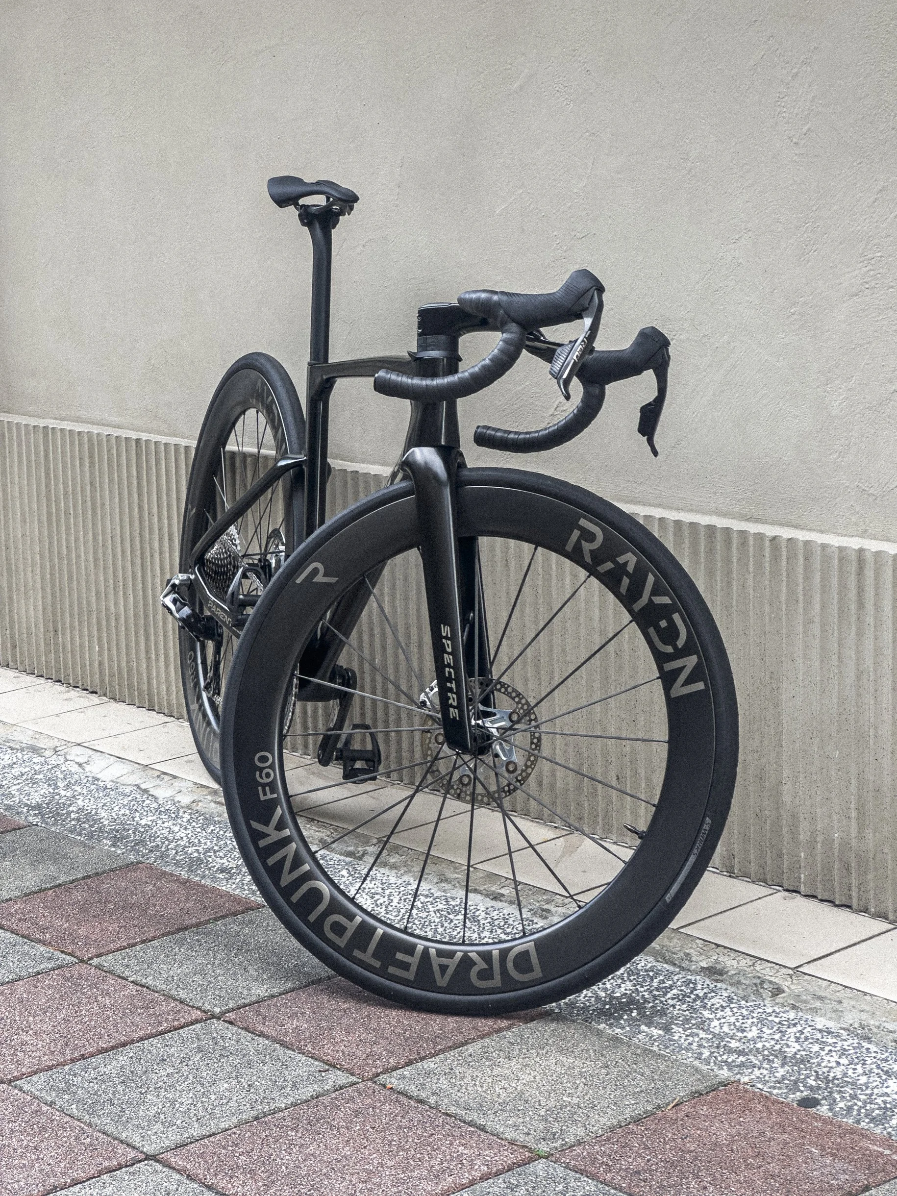 A PARENTESI  black road bike with deep-section RAYDN wheels, parked against a beige wall on a sidewalk.