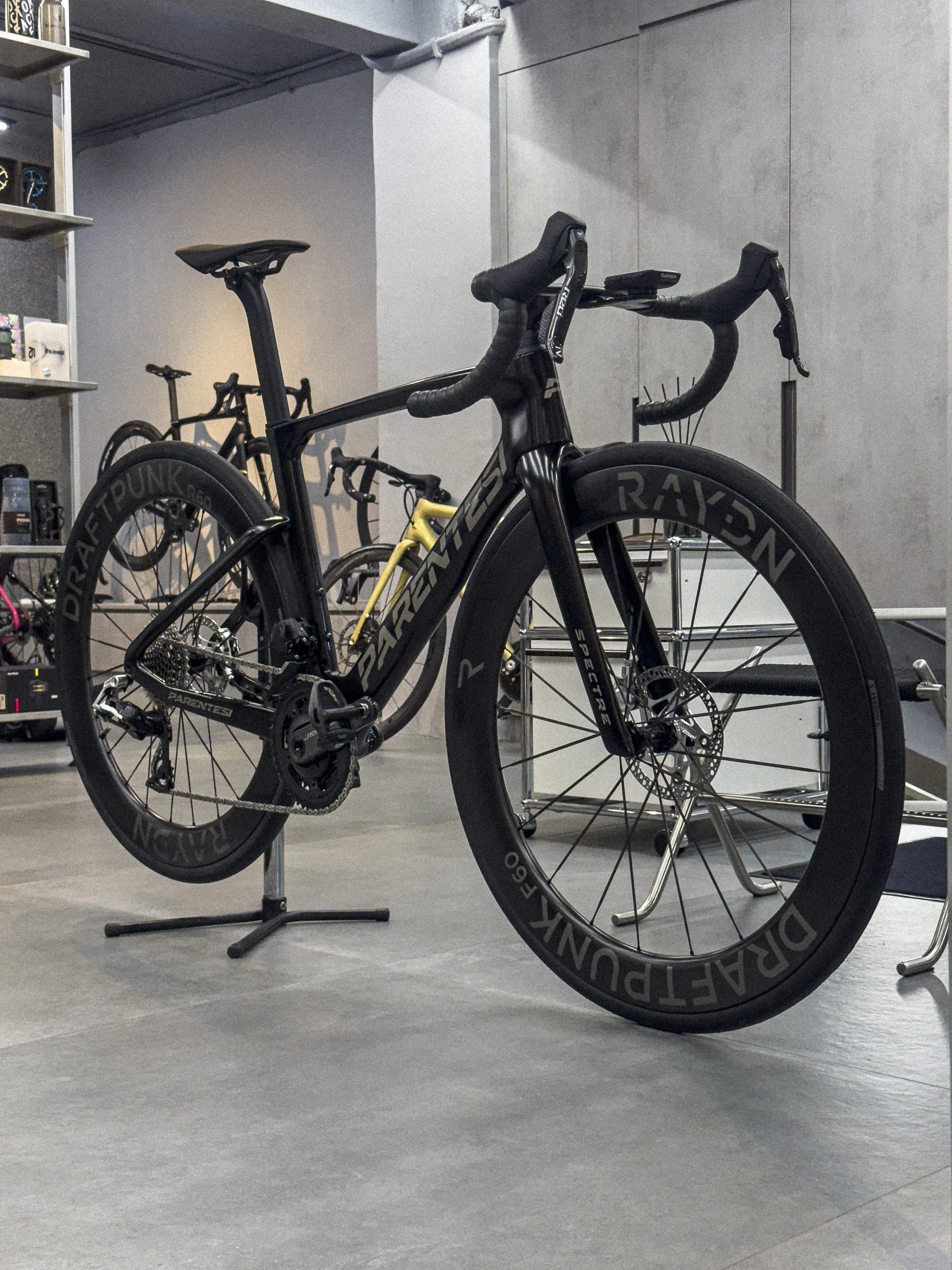 A black PARENTESI bicycle with race wheels and disc brakes is mounted on a repair stand in a bicycle shop, with other bikes and gear in the background.