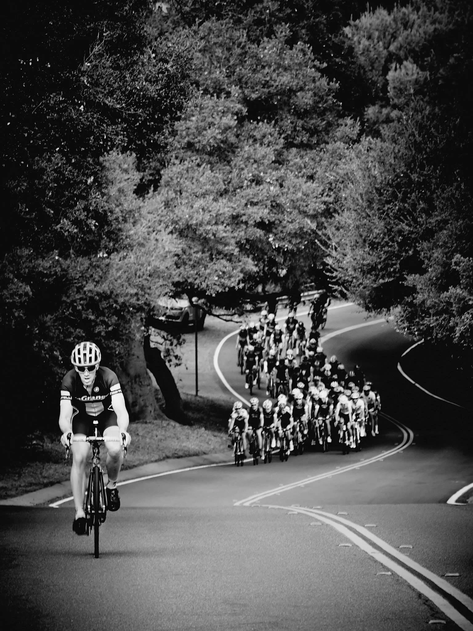A cyclist ahead of a large group of cyclists riding on a winding road surrounded by trees.