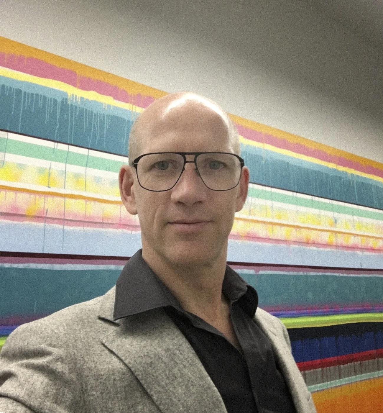 A man with glasses, wearing a beige blazer and black shirt, taking a selfie in front of an abstract colorful painting with horizontal stripes of various colors and textures.