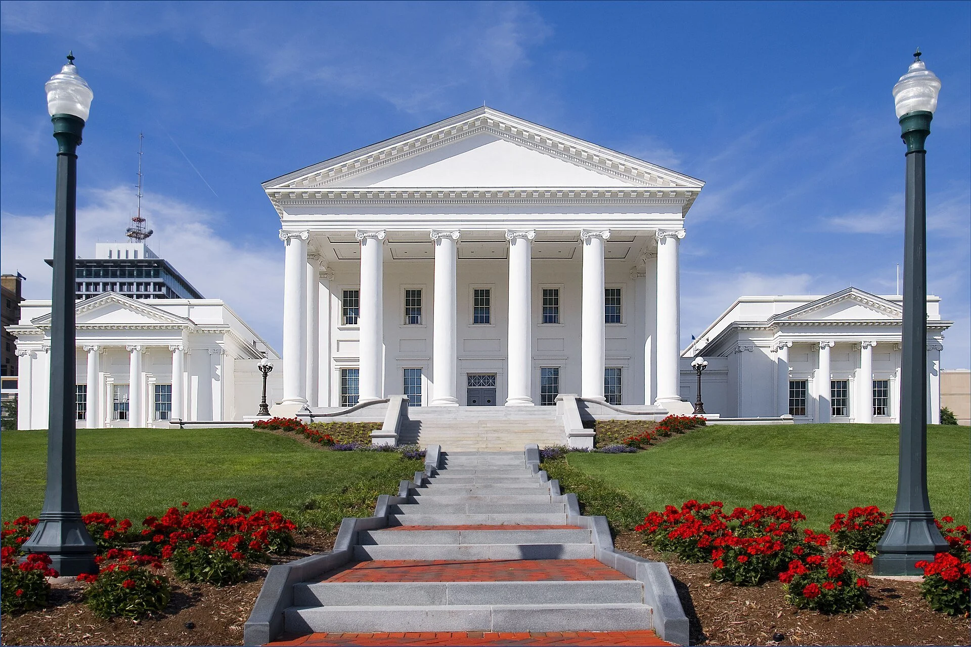 The Virginia State Capitol in Richmond, a white temple-style building with columns, behind red flower beds and lampposts.
