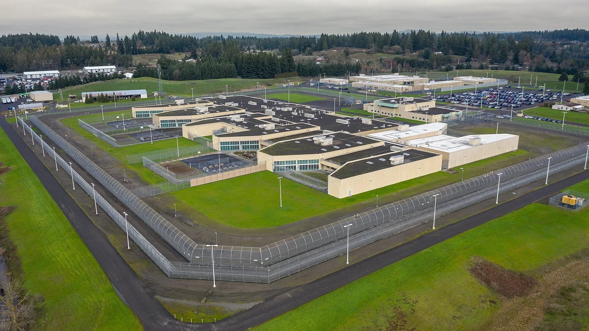 An aerial view of Coffee Creek Correctional Facility in Wilsonville, Oregon: tan, flat-roofed buildings inside a double perimeter fence topped with coiled wire, surrounded by green lawns, with a parking lot, more buildings, and forested hills under a