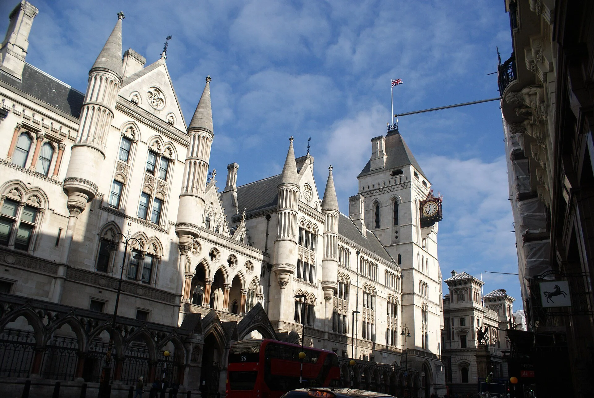 The Royal Courts of Justice on the Strand in London, a white Gothic Revival building with pointed towers, arched windows, and a clock, under a blue sky, with a red double-decker bus passing in front.