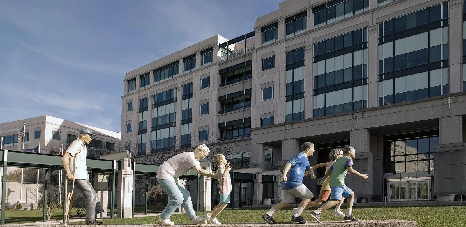 A modern multistory office building with stone walls and rows of dark windows under a blue sky. In front, a row of painted sculptures of figures walks and runs across the lawn, from an older figure with a crutch to several children.