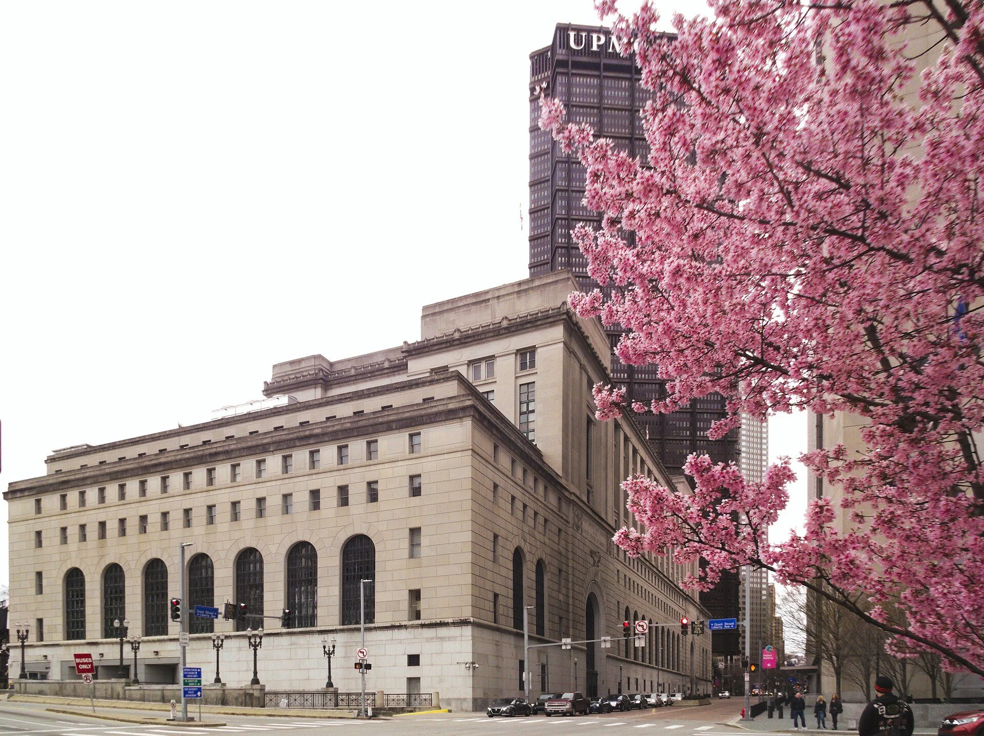 A large beige stone federal courthouse with tall arched windows on a downtown Pittsburgh corner, a dark skyscraper topped with UPMC letters behind it, and a tree in pink bloom in the foreground.