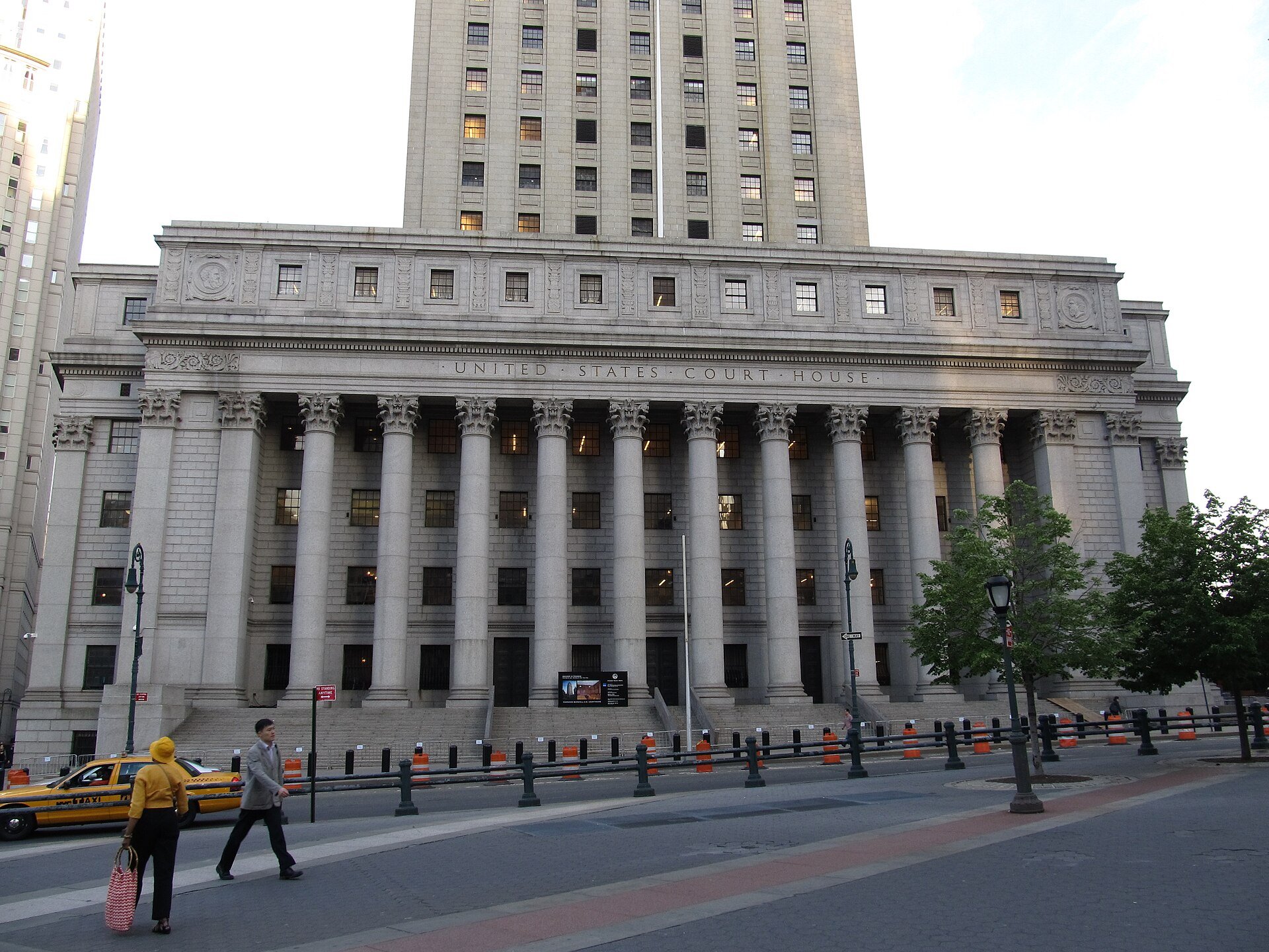 The Thurgood Marshall U.S. Courthouse in Lower Manhattan, a gray granite building with a long row of tall columns under the carved words United States Court House and a tower rising behind it. Two people walk across the plaza in front as a yellow tax
