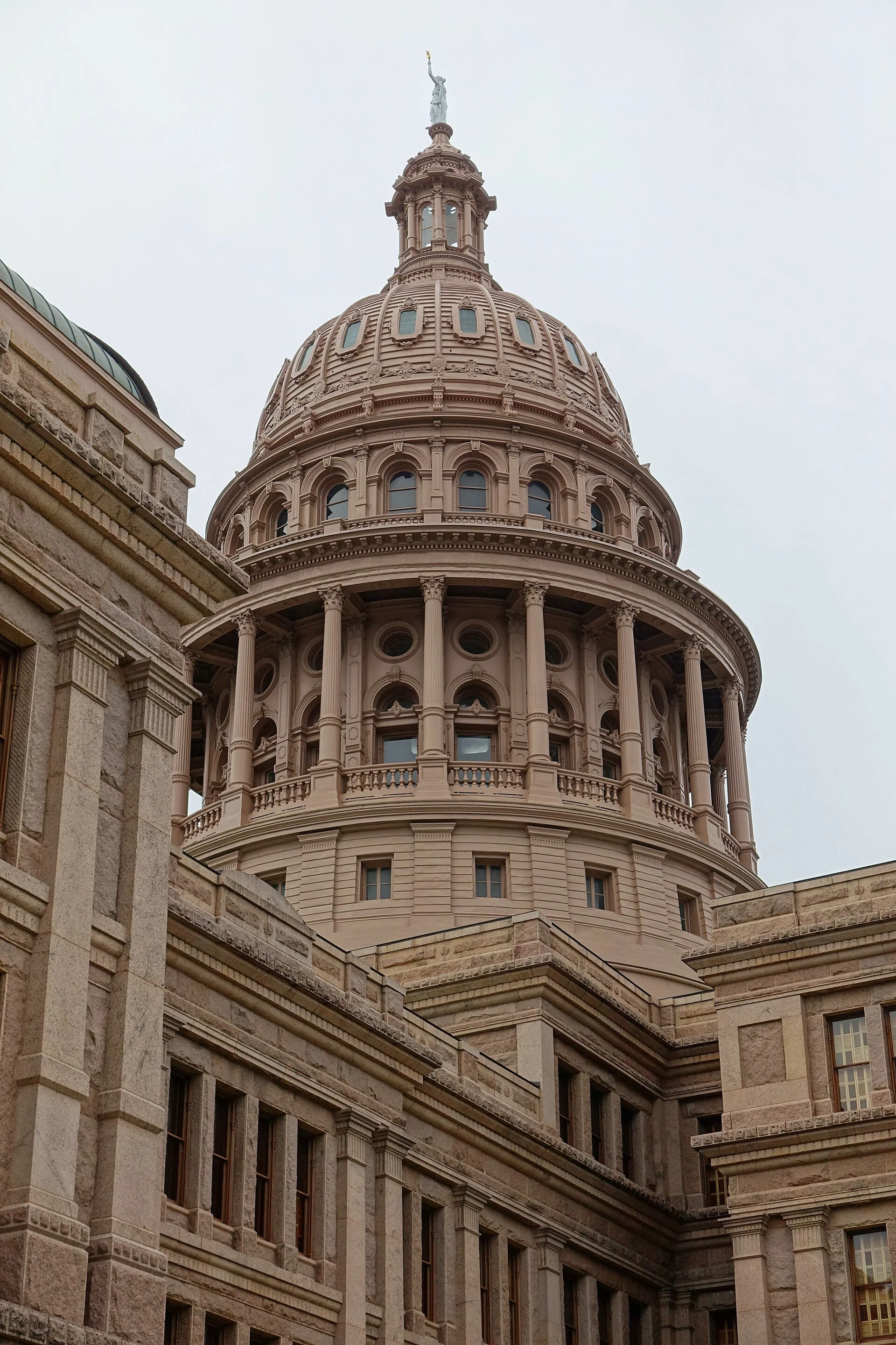 The dome of the Texas State Capitol in Austin, topped by a statue, rising above pink granite walls against a pale sky.