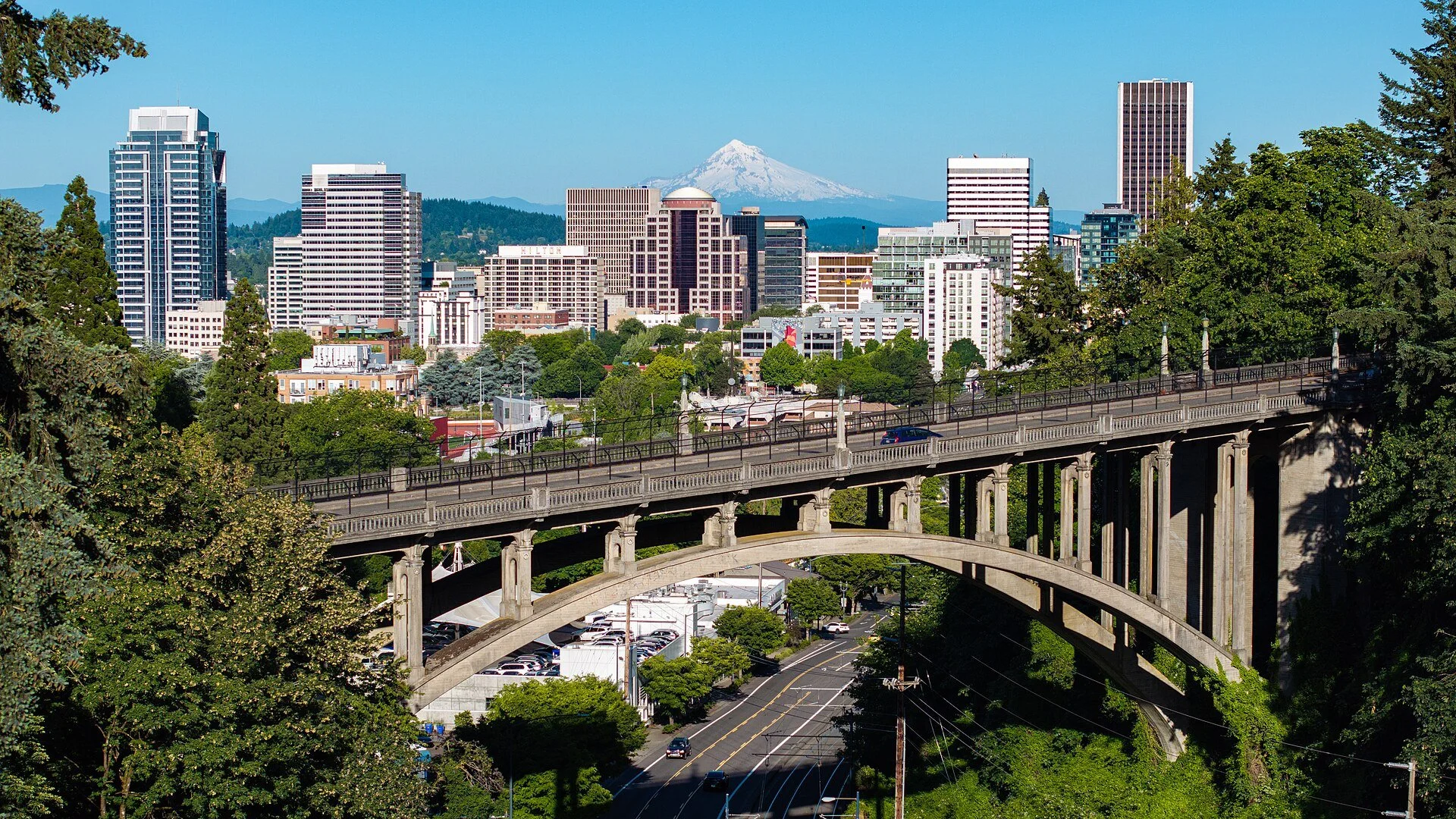 Downtown Portland's towers rise behind the arched Vista Bridge, with snow-capped Mount Hood on the horizon under a clear blue sky.