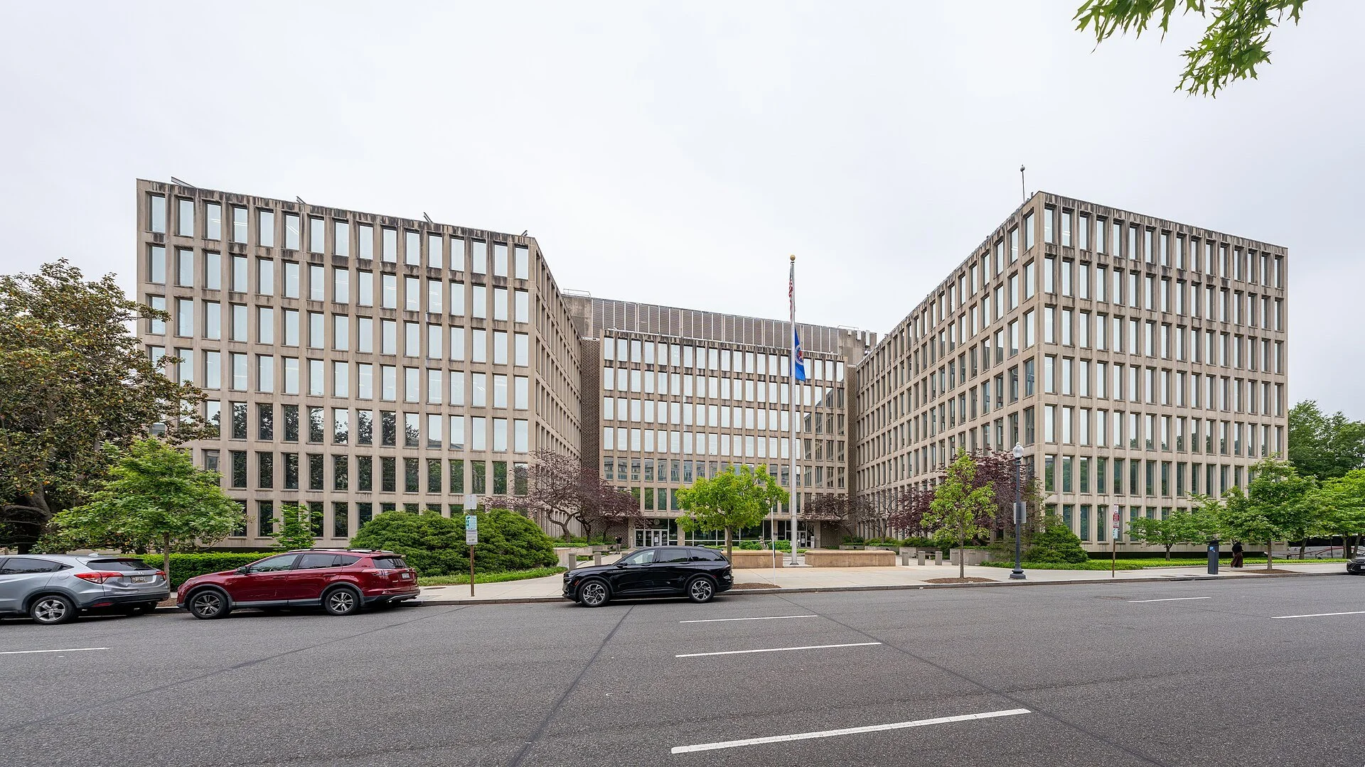 The Theodore Roosevelt Federal Building in Washington, D.C., headquarters of the U.S. Office of Personnel Management, a pale concrete office building with a grid of tall windows, a flagpole out front, and cars parked along the street.