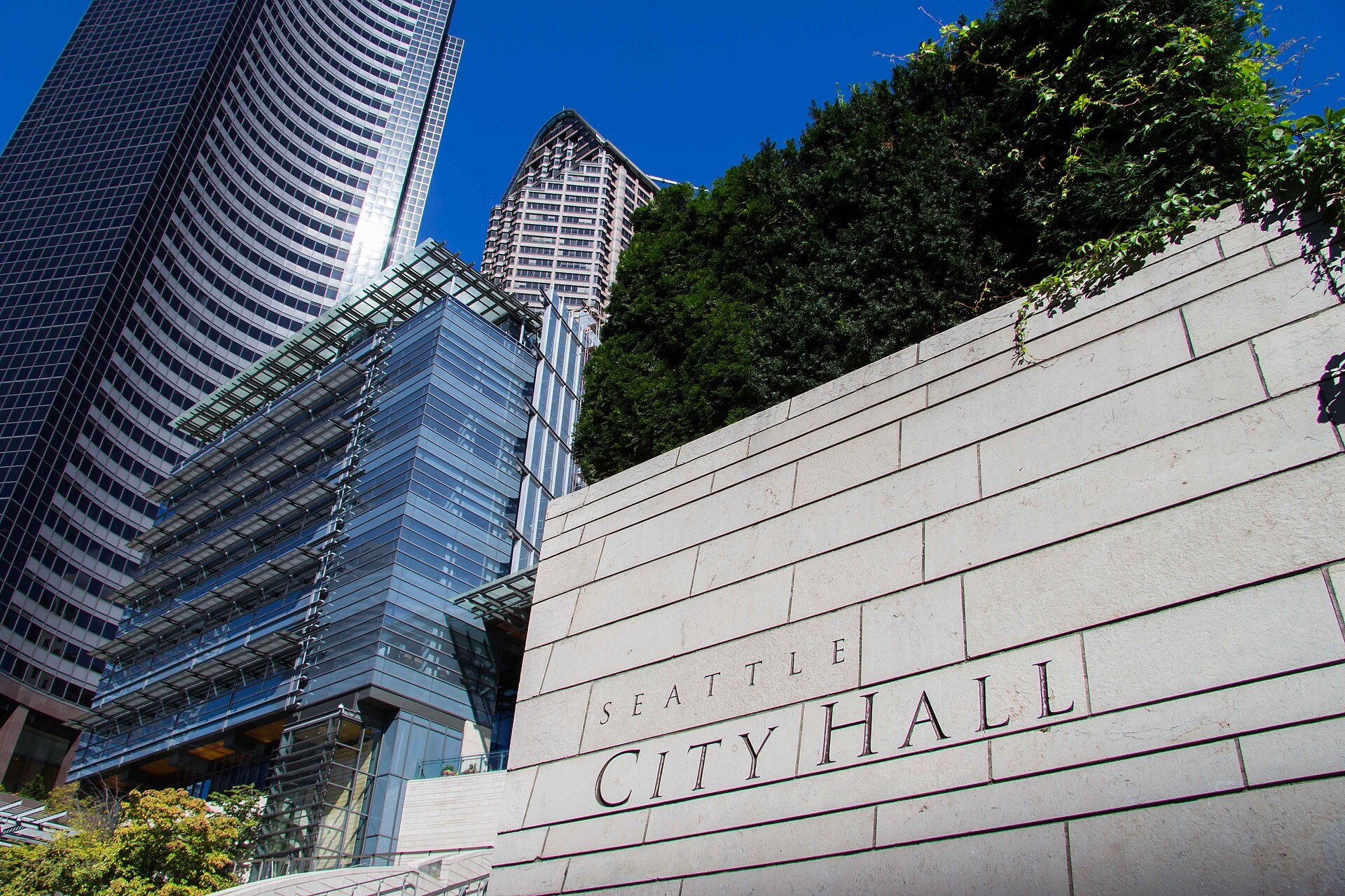 A pale stone wall engraved with the words Seattle City Hall, with the glass and steel City Hall building and taller downtown towers rising behind it under a deep blue sky.