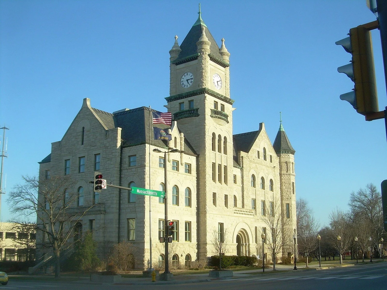 A historic limestone courthouse with a clock tower, turrets, and arched windows on a clear day, with U.S. and Kansas flags out front and a Massachusetts Street sign at the corner.