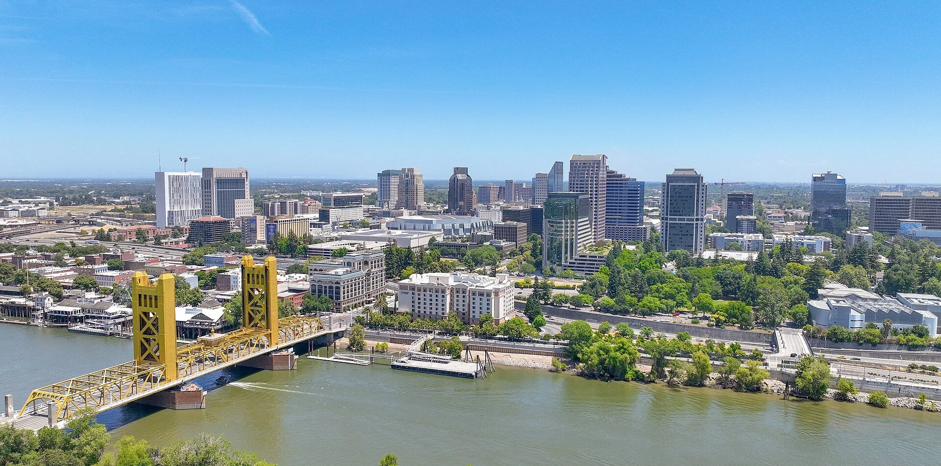 Downtown Sacramento seen from above on a clear day, with the gold-colored Tower Bridge crossing the Sacramento River in the foreground and a cluster of office towers behind it.