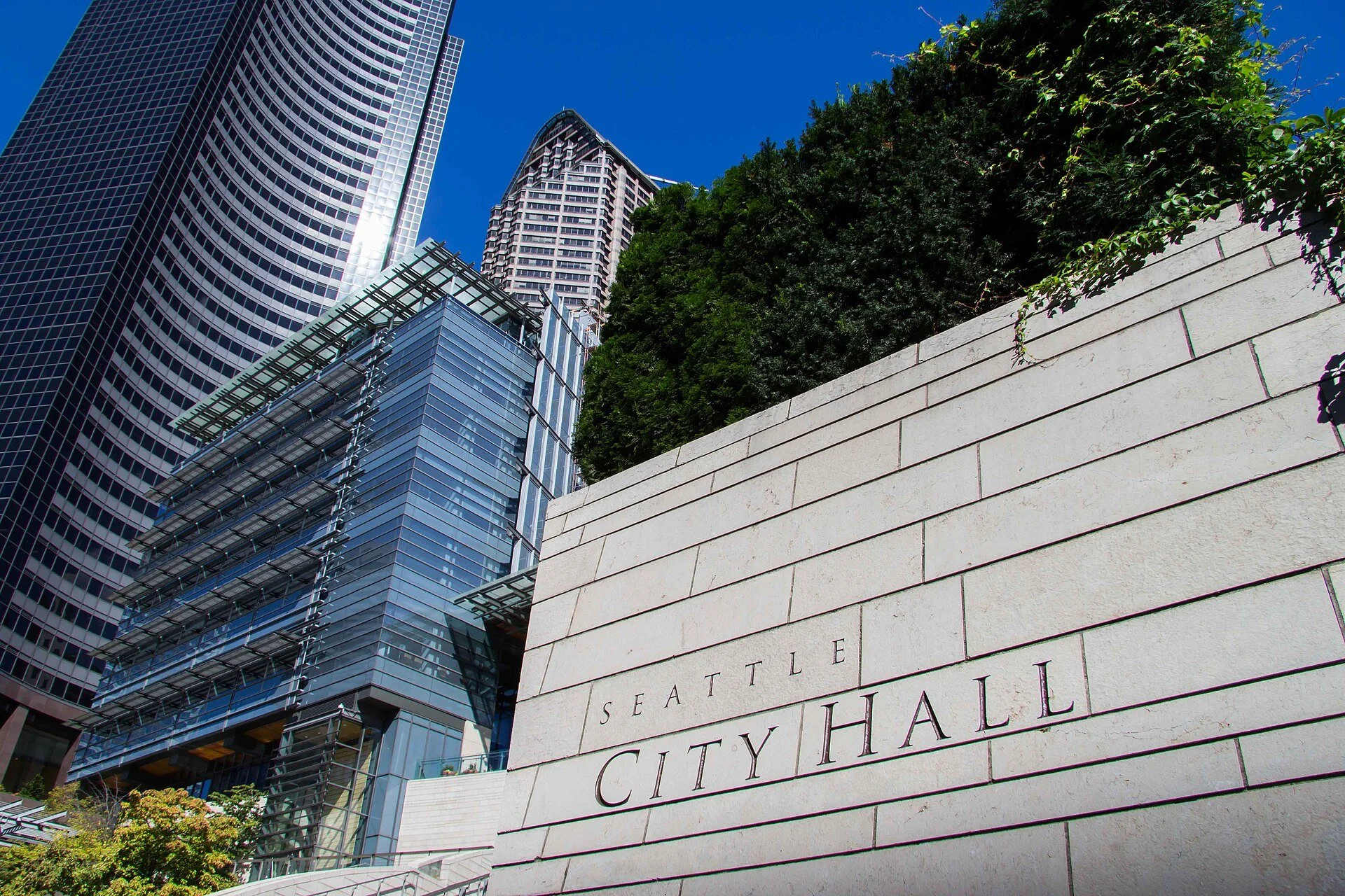 Carved stone sign reading Seattle City Hall in front of a glass civic building and downtown towers against a blue sky.