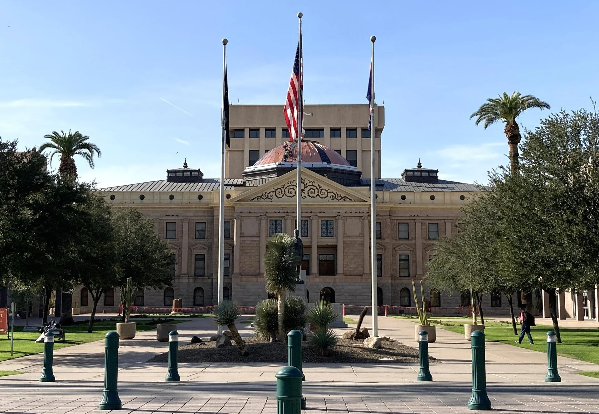 The Arizona State Capitol in Phoenix, a stone building with a copper dome, behind flagpoles, palm trees and a landscaped plaza on a sunny day.