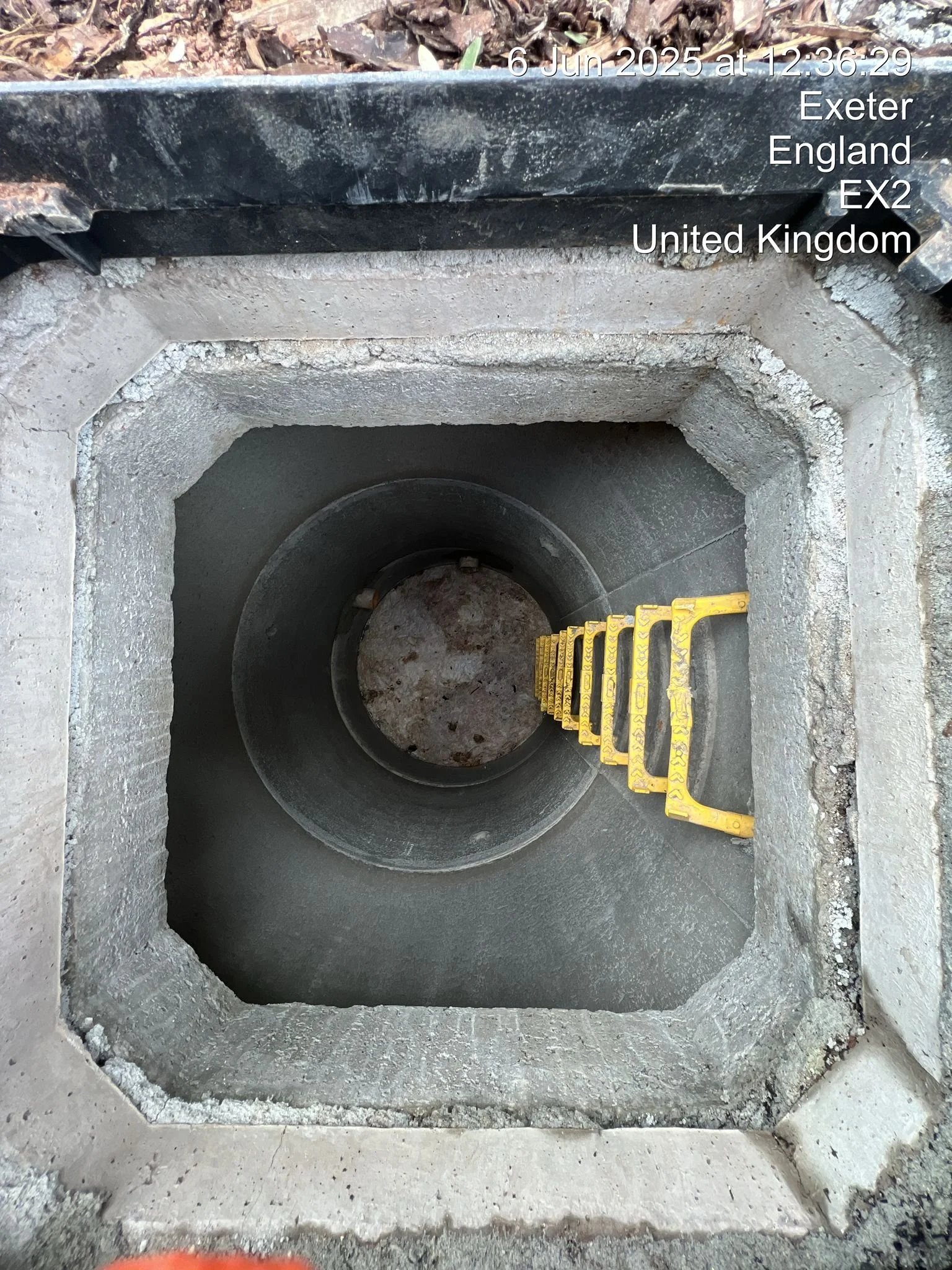 View looking down into a street inspection hole with a yellow measuring tool inside, surrounded by concrete and earth, with a timestamp and location overlay.