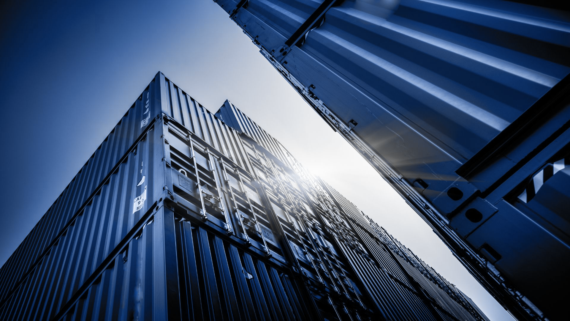 Blick nach oben auf gewölbte Container aus Metall, die in verschiedenen Richtungen gestapelt sind, mit der Sonne im Hintergrund.