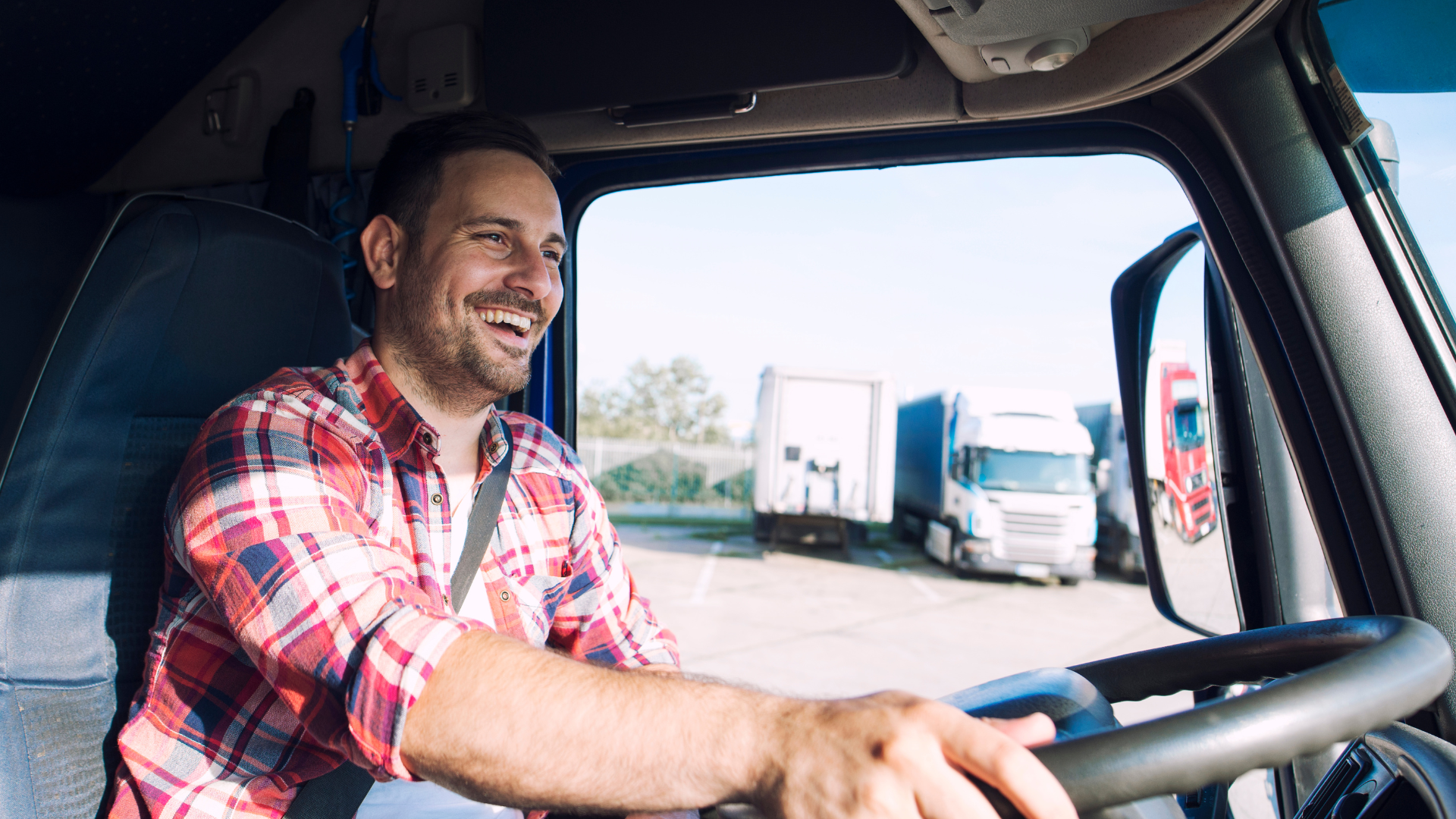 Lächelnder LKW-Fahrer im Fahrerraum bei Tageslicht, mit mehreren LKWs im Hintergrund auf einem Parkplatz.