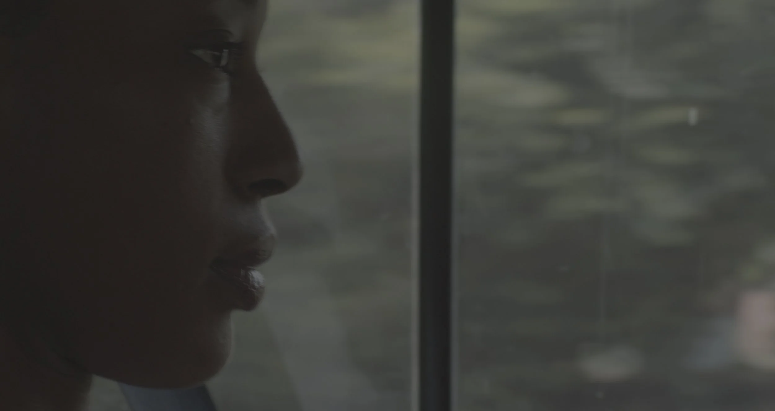 Close-up of a woman's face in profile, with focus on her lips and nose, as she looks through a glass window or divider, with blurred greenery in the background.