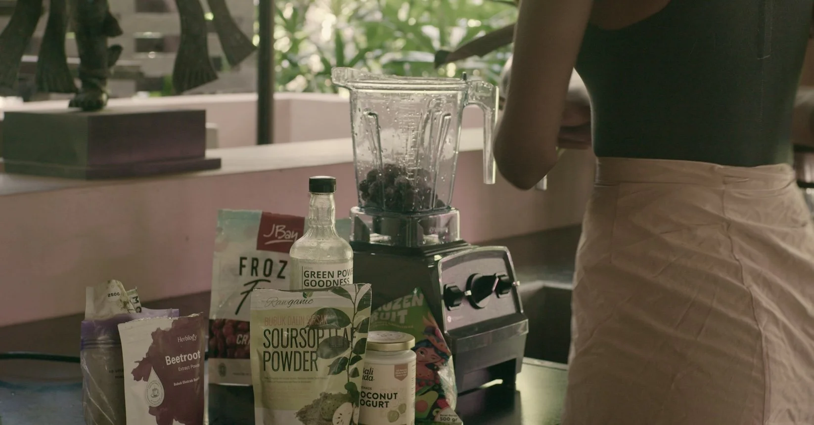 A person making a smoothie using a blender filled with dark berries. The counter holds various health food ingredients including beetroot powder, sour sop leaf powder, coconut yogurt, and a bottle of green powder. Sunlight filters through greenery outside.