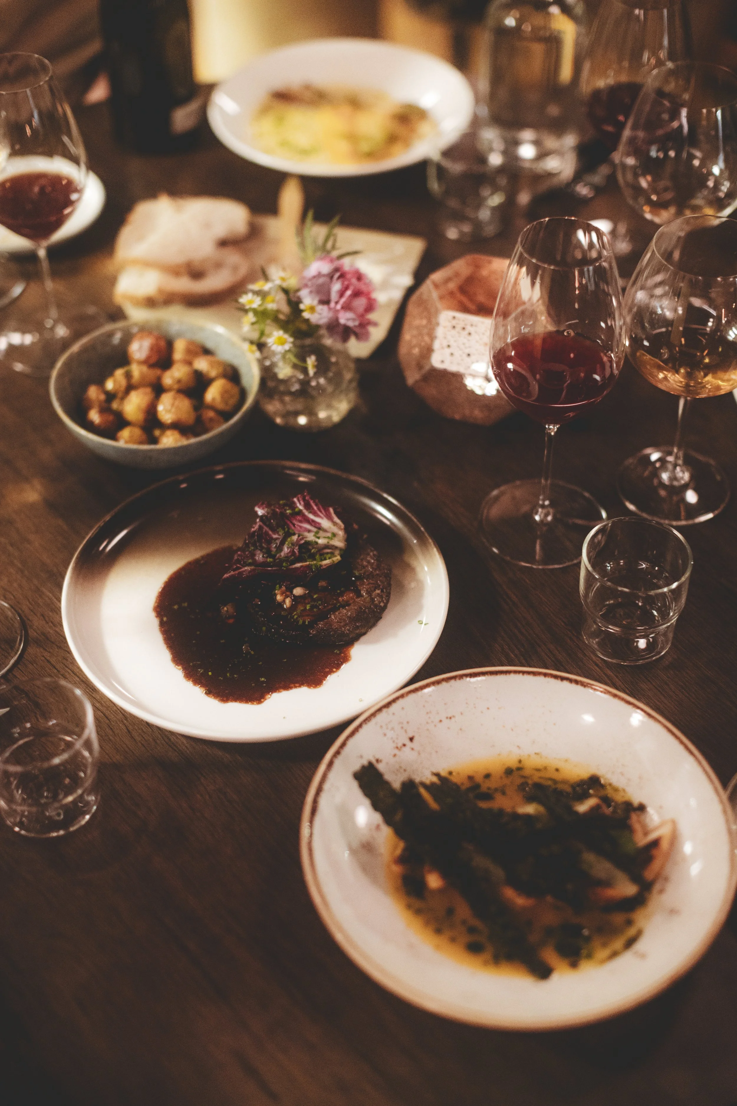 Et bord med flere glass med rødvin, et fat med en biff i saus, en tallerken med sjømat, og en liten blomsterbukett.