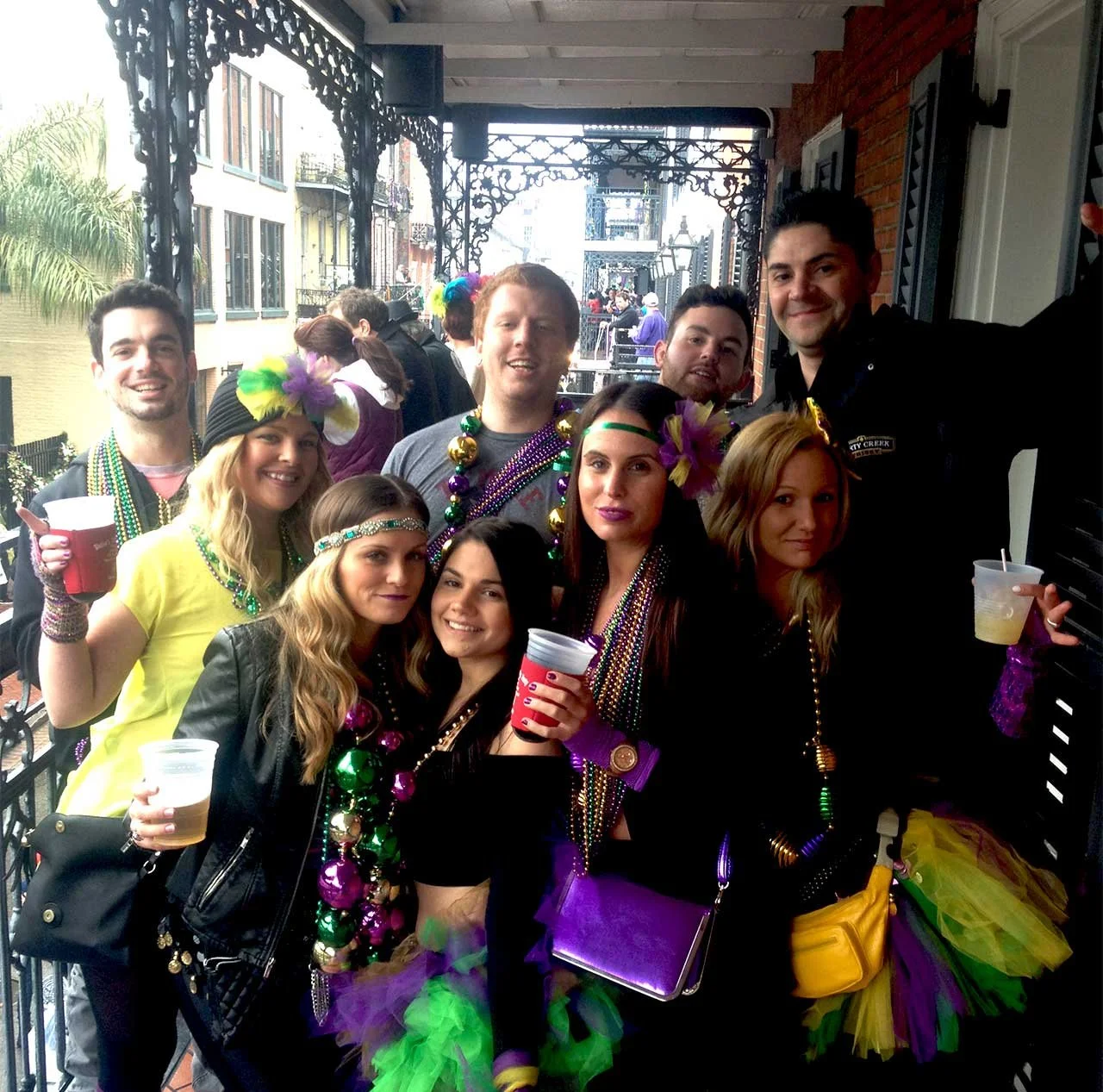 Group of people celebrating Mardi Gras on a balcony, wearing colorful beads, feathered masks, and festive costumes, holding drinks.
