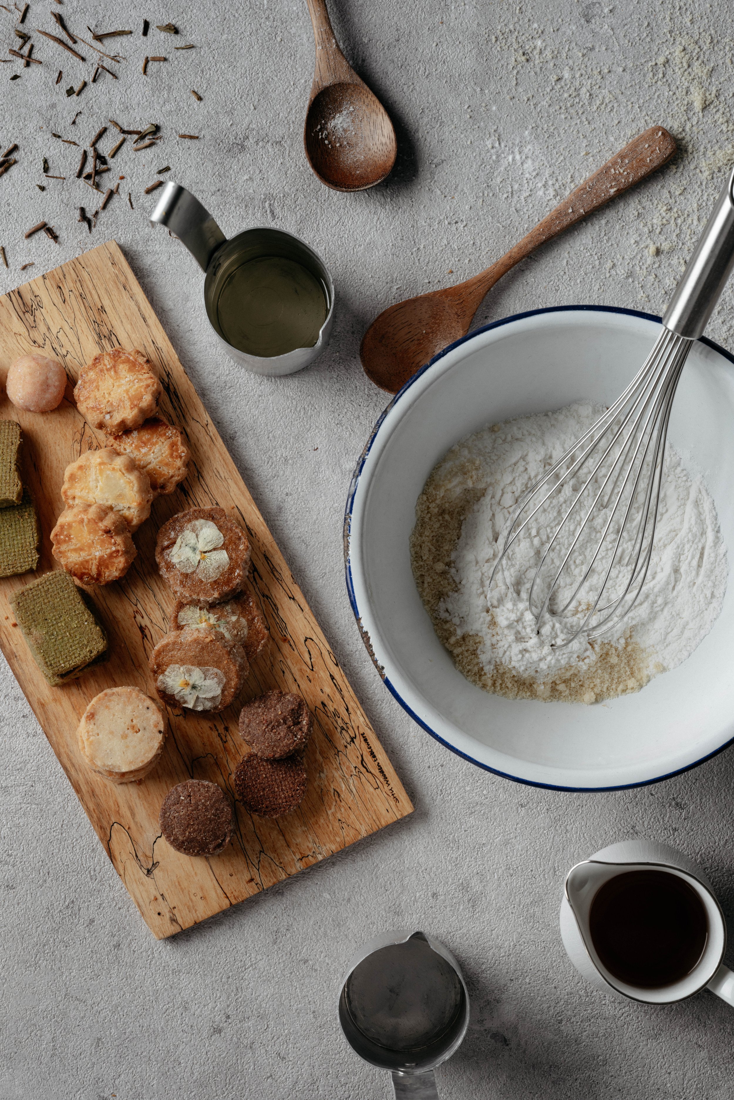 粉と砂糖などの材料を混ぜるためのホイッパーとボウル、クッキー型や乾燥したクッキー、ケーキの材料が置かれた木製のトレーがテーブルに並んでいるキッチンのシーン