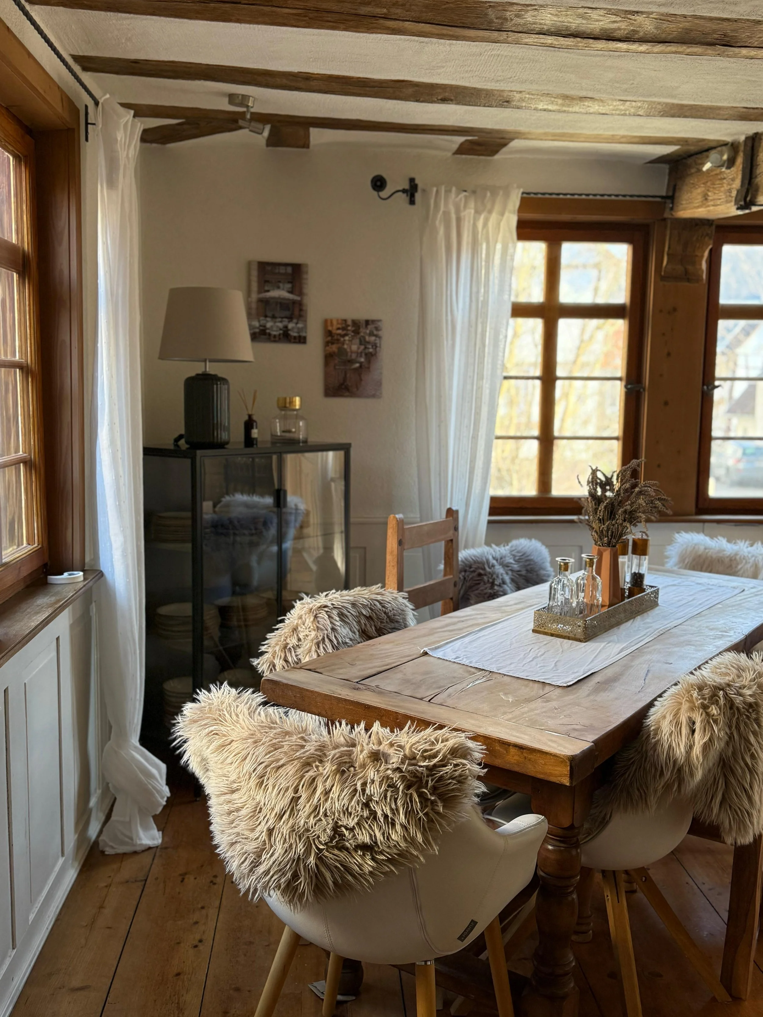 Gemütlicher Essbereich mit Holztisch, Samtstühlen mit pelzigen Überzügen, Fenstern und Weißvorhängen in einem rustikalen Zimmer.
