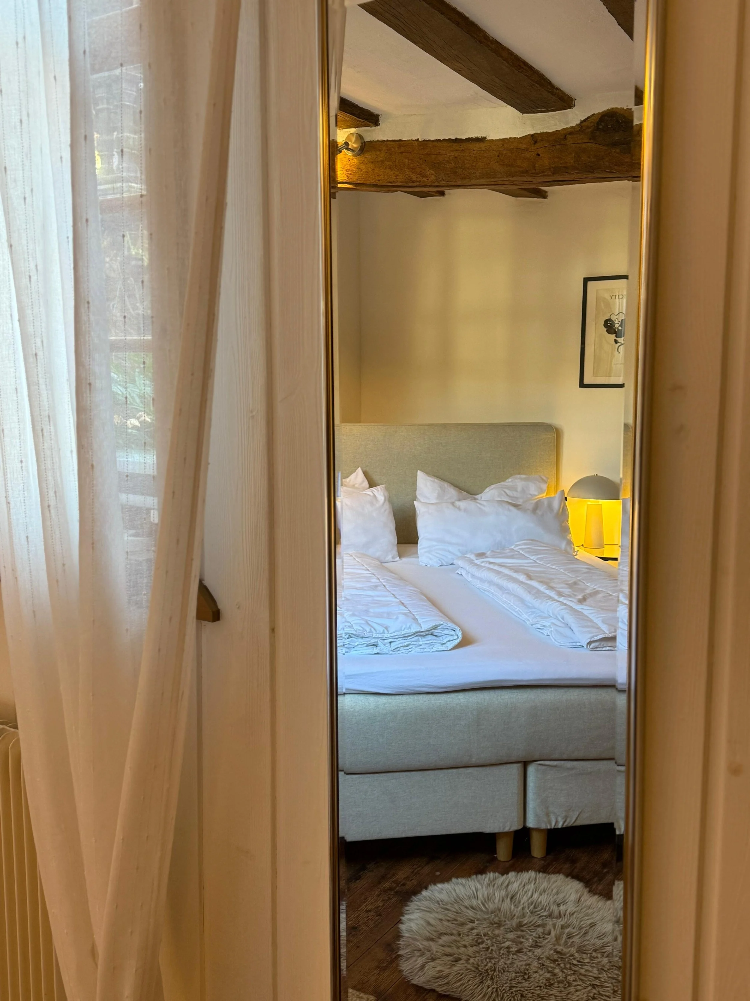 Ein Blick in ein Schlafzimmer durch einen Spiegel, zeigt ein Bett mit weißen Bettwäsche, ein Nachttisch mit einer Lampe, eine Wand mit einem Bild und eine Decke aus Holzbalken.