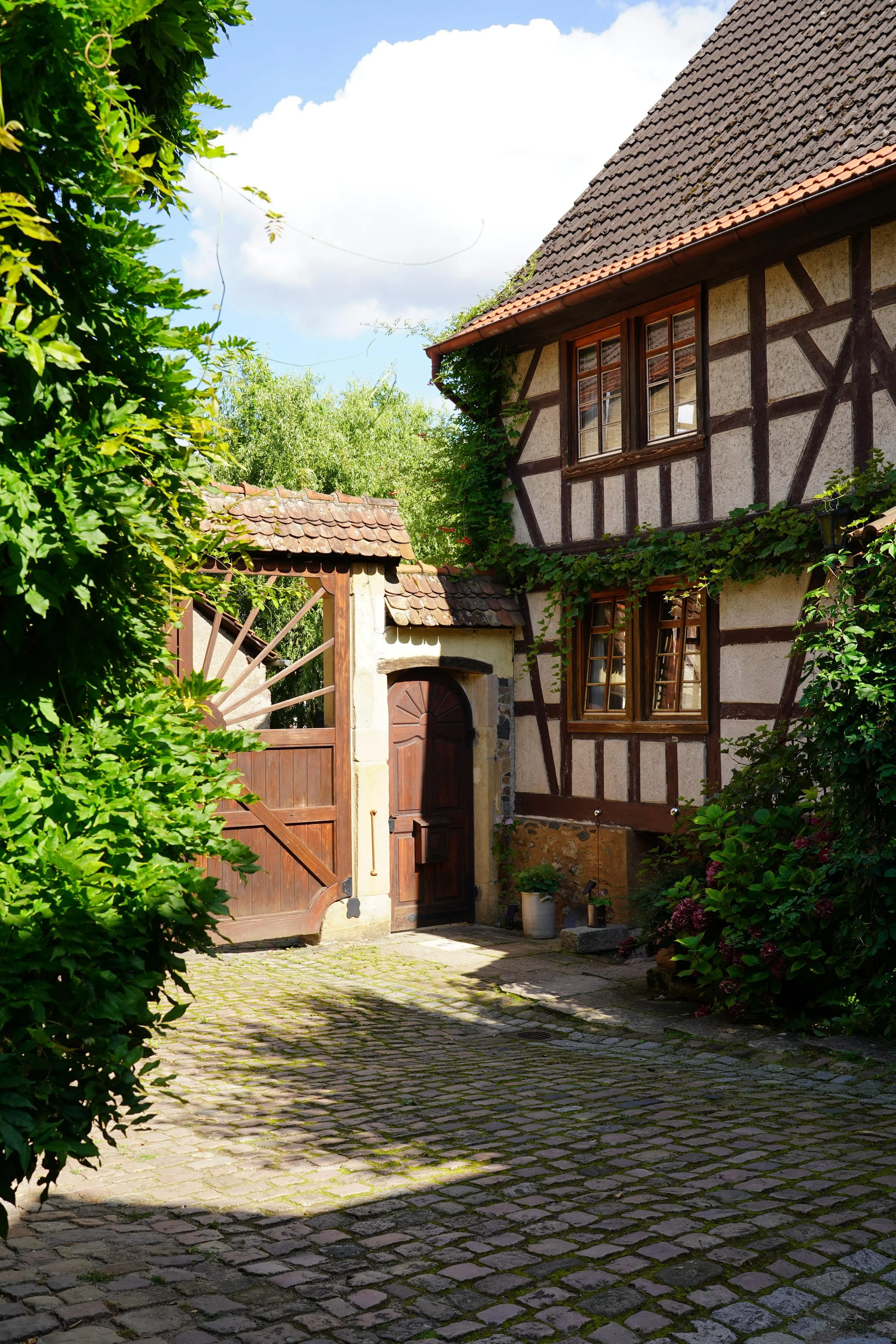 Ein malerisches Fachwerkhaus mit Holzrahmen, umgeben von grünen Pflanzen und einem gepflasterten Hof unter einem blauen Himmel mit Wolken.