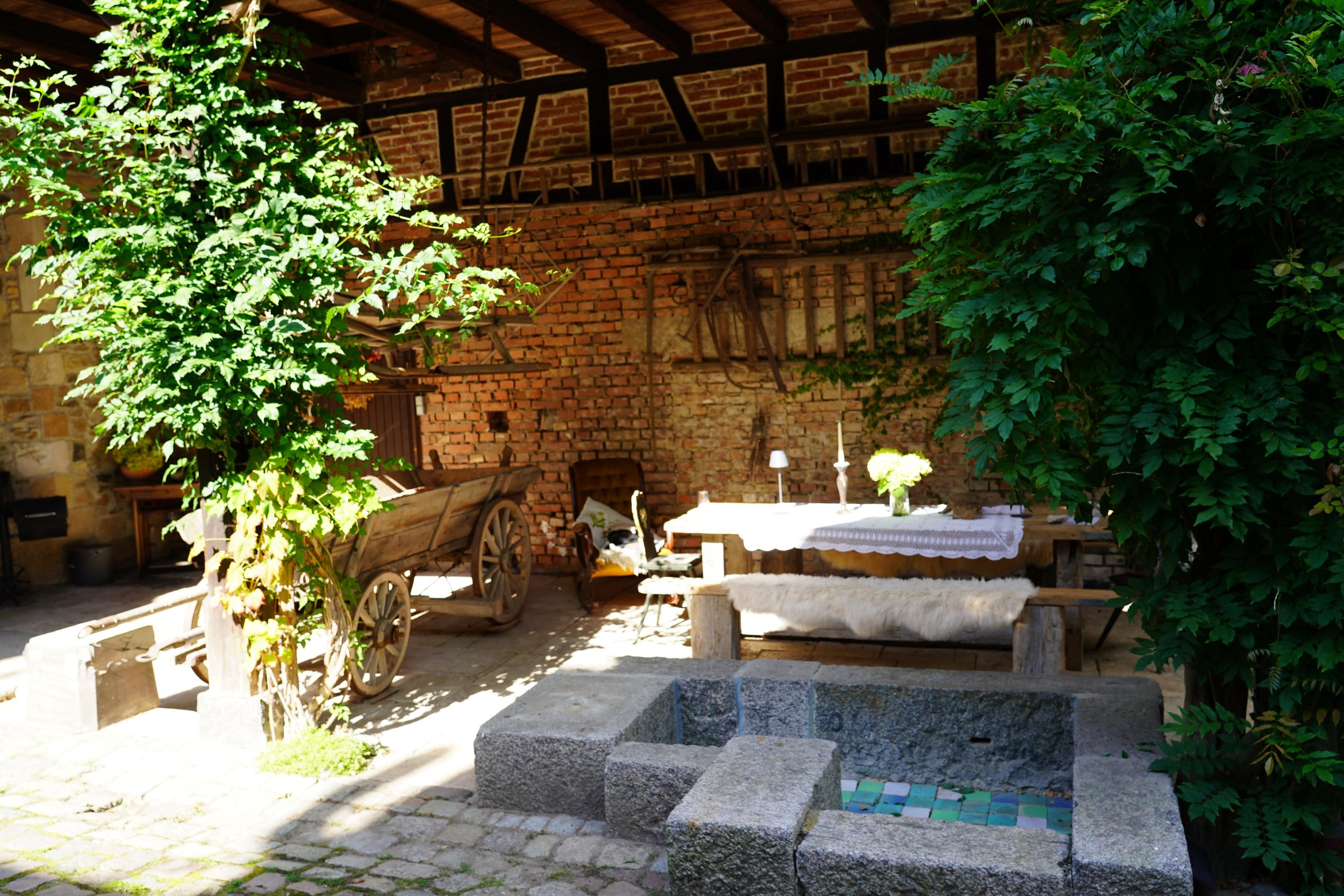 Ein gemütlicher, rustikaler Innenhof mit einer alten Holzkarre, einem hölzernen Tisch mit weißer Tischdecke, Kerzen, einem Blumentopf und einem bequemen Stuhl. Der Raum ist von Ziegelwänden und grünen Bäumen umgeben.