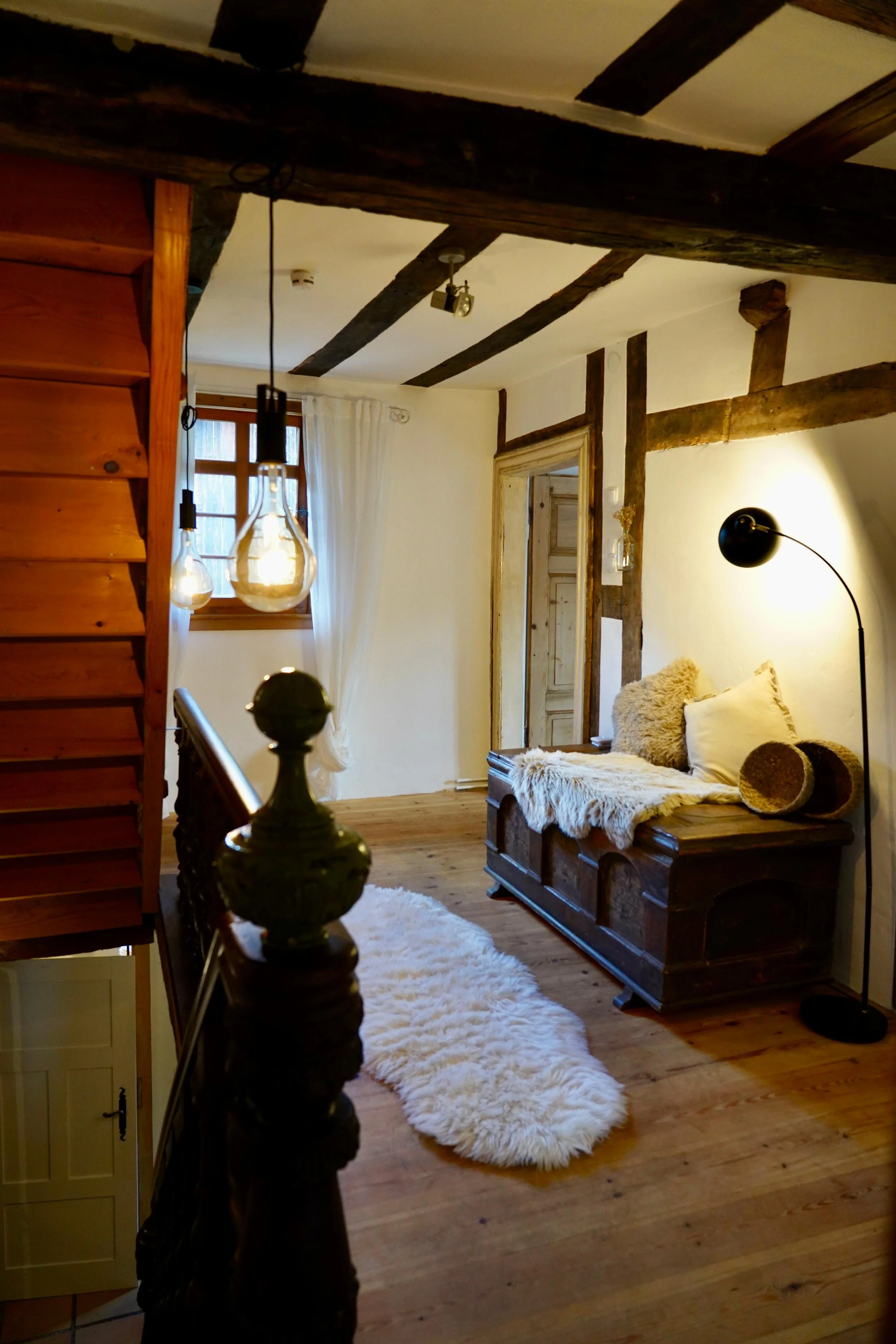 Gemütliches Zimmer mit Holzdecke, einem Fenster mit weißen Vorhängen, einer schwarzen Stehlampe, einer Sitztruhe mit Kissen und Fell, Holzfußboden und Teppichlien.