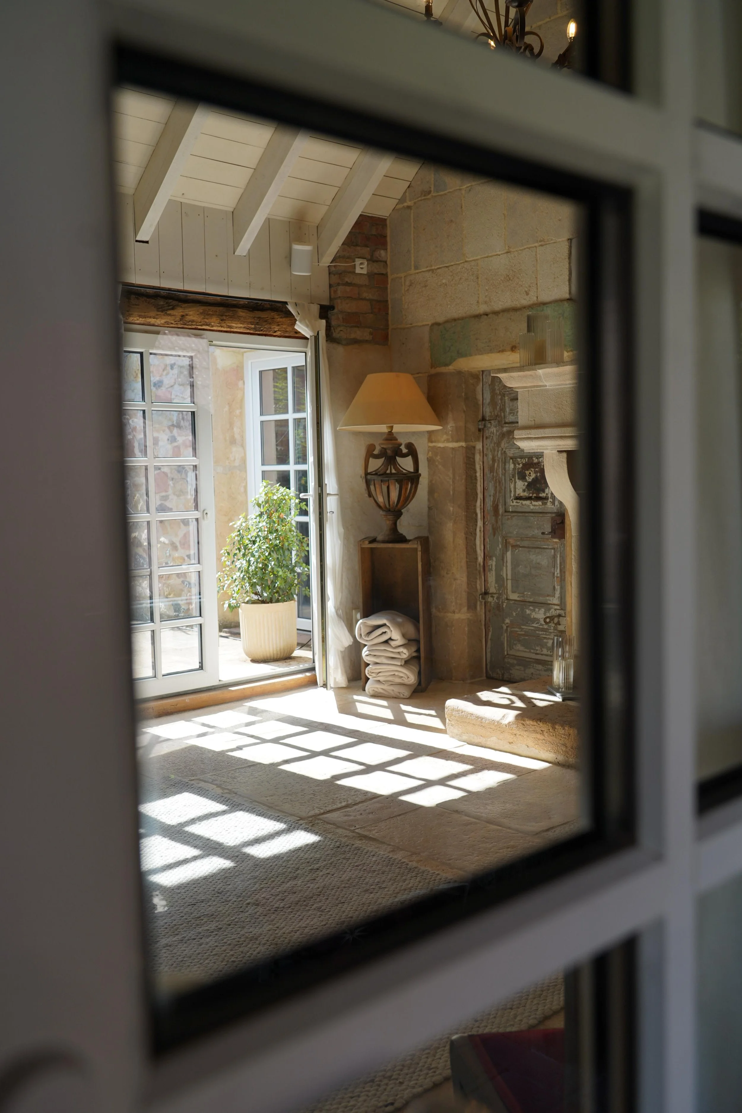 Ein heller Raum mit einer offenen Terrassentür, die Sonnenschein und Schatten auf den Boden wirft. Im Raum befindet sich ein Kamin, eine Stehlampe, ein Regal mit gefalteten Handtüchern und eine Pflanze auf der Terrasse im Hintergrund.