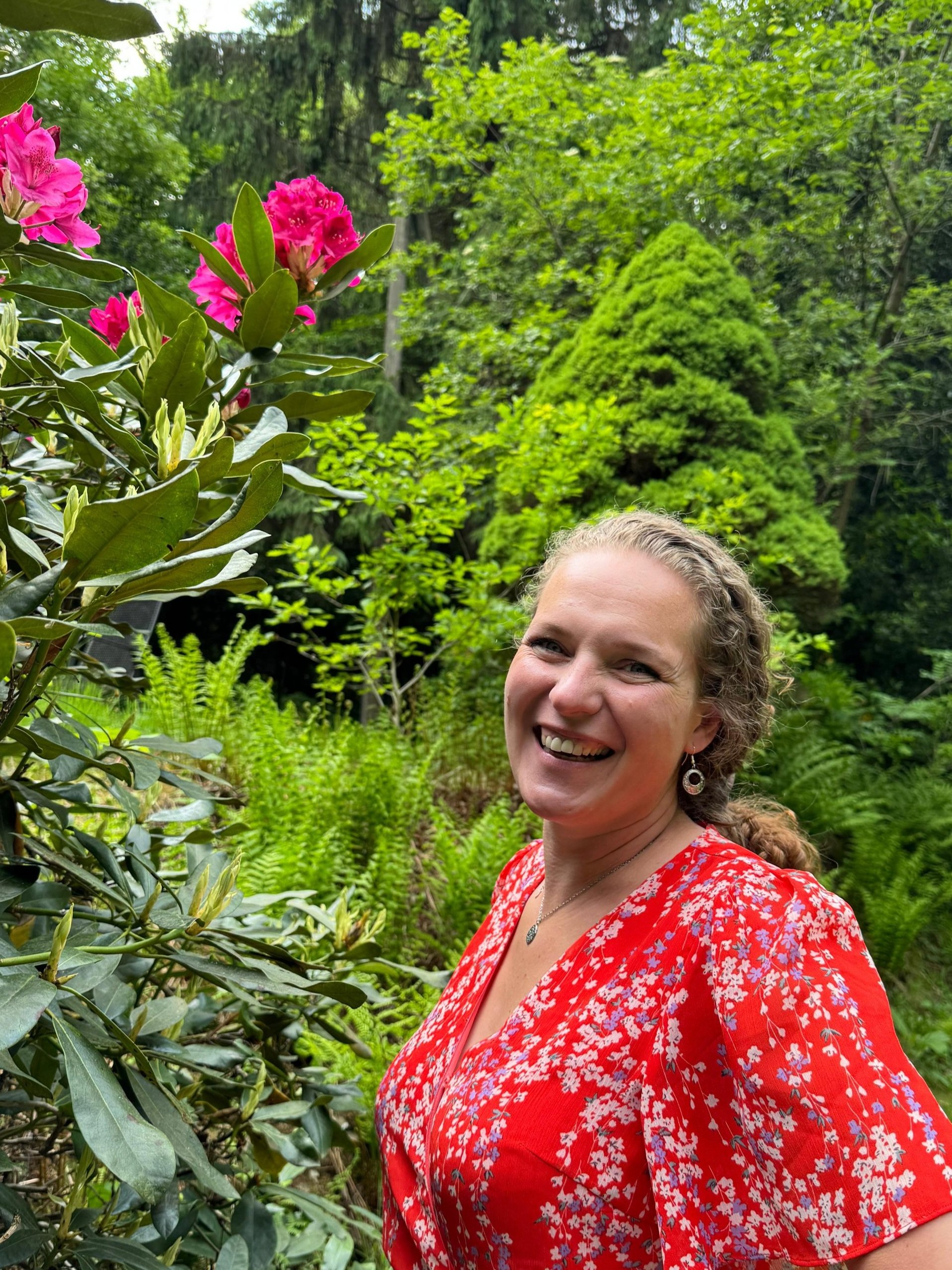 Eine lachende Frau in einem roten, blumigen Kleid im Garten mit grünen Büschen und Bäumen.