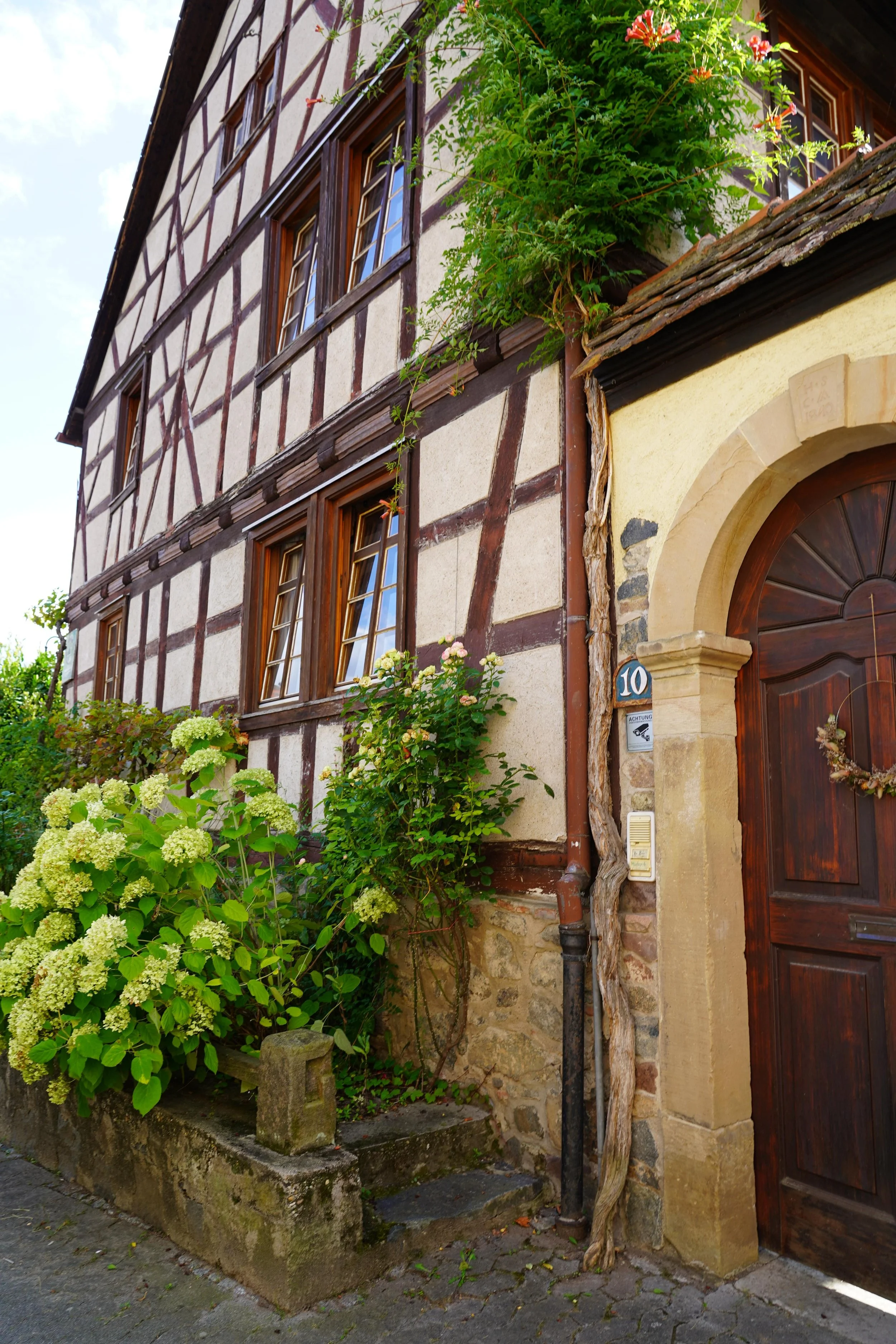 Traditionelles Fachwerkhaus mit Blumen im Fenster und Vegetation vor der Tür, Haustür mit Kranz, Hausnummer 10.