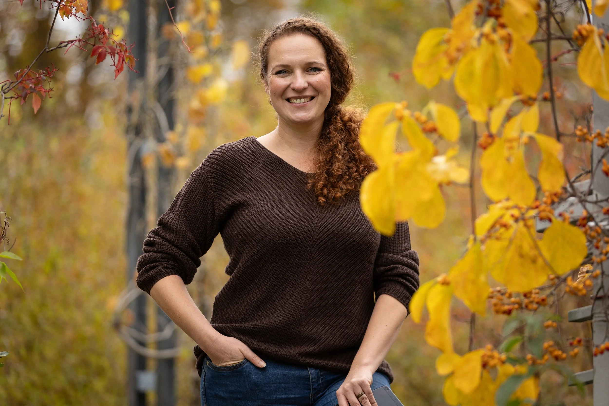 Junge Frau mit braunem Pullover, lächelnd, stehend in einem herbstlichen Park, umgeben von gelben und roten Blättern.