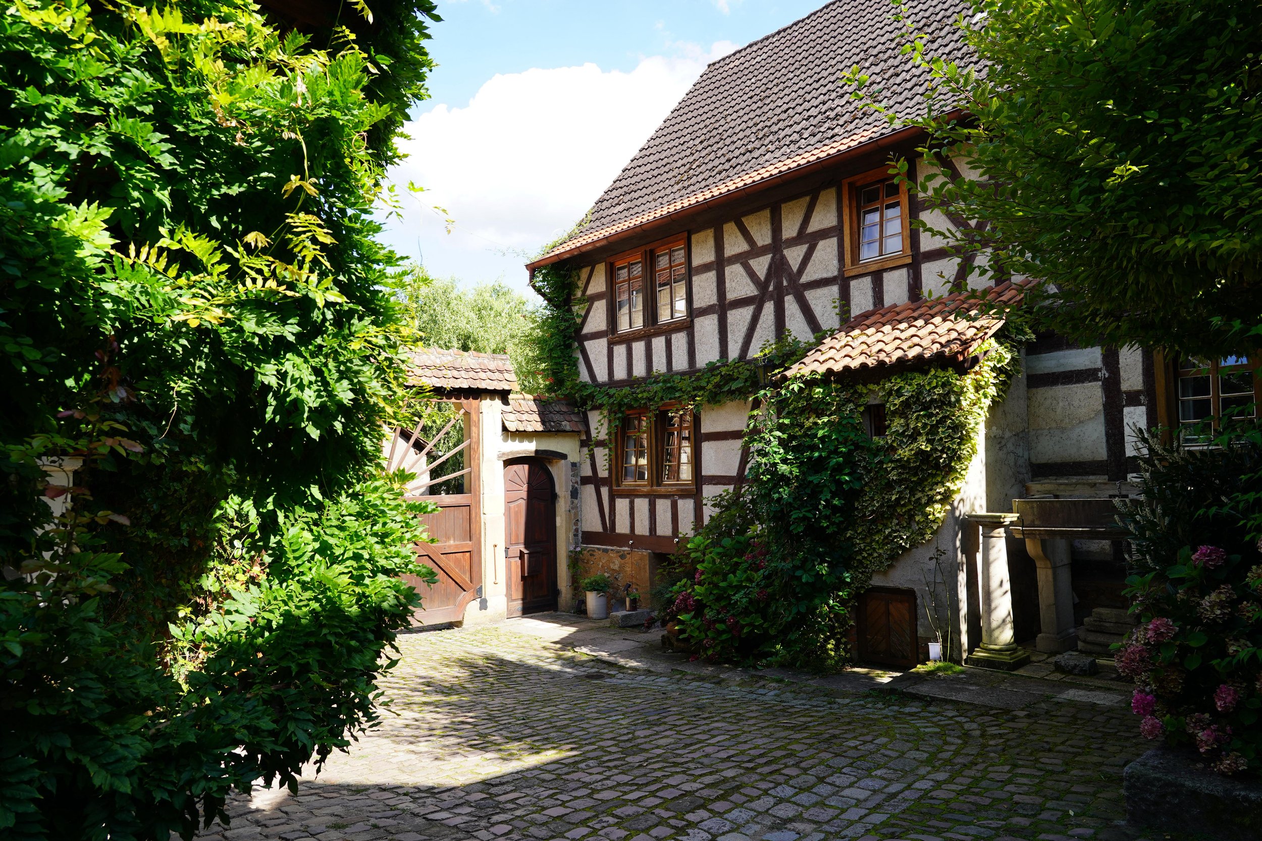 Ein traditionelles, Fachwerkhaus mit grünen Pflanzen an den Wänden und einem Holztor im Hof, umgeben von Bäumen und Blumen, bei Tageslicht